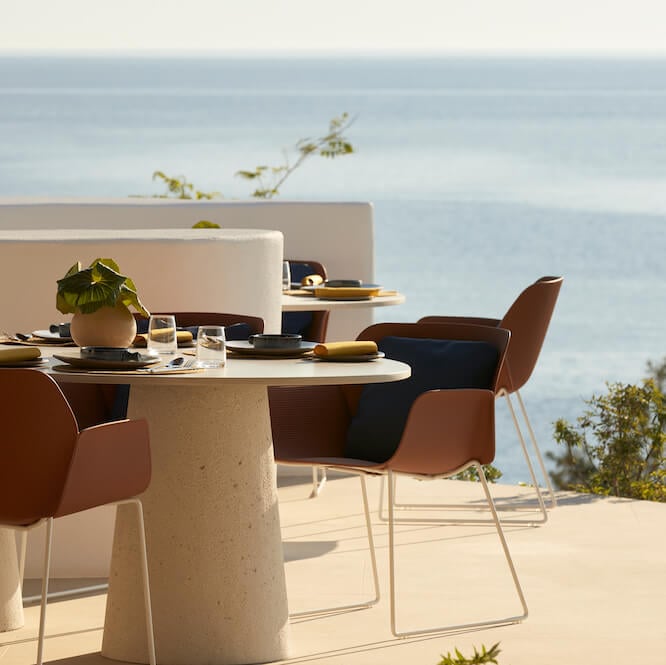 Pārocks Restaurant, Europe — Coastal dining terrace with round travertine table, cognac leather chairs, sea views, and minimalist modern design in natural light.