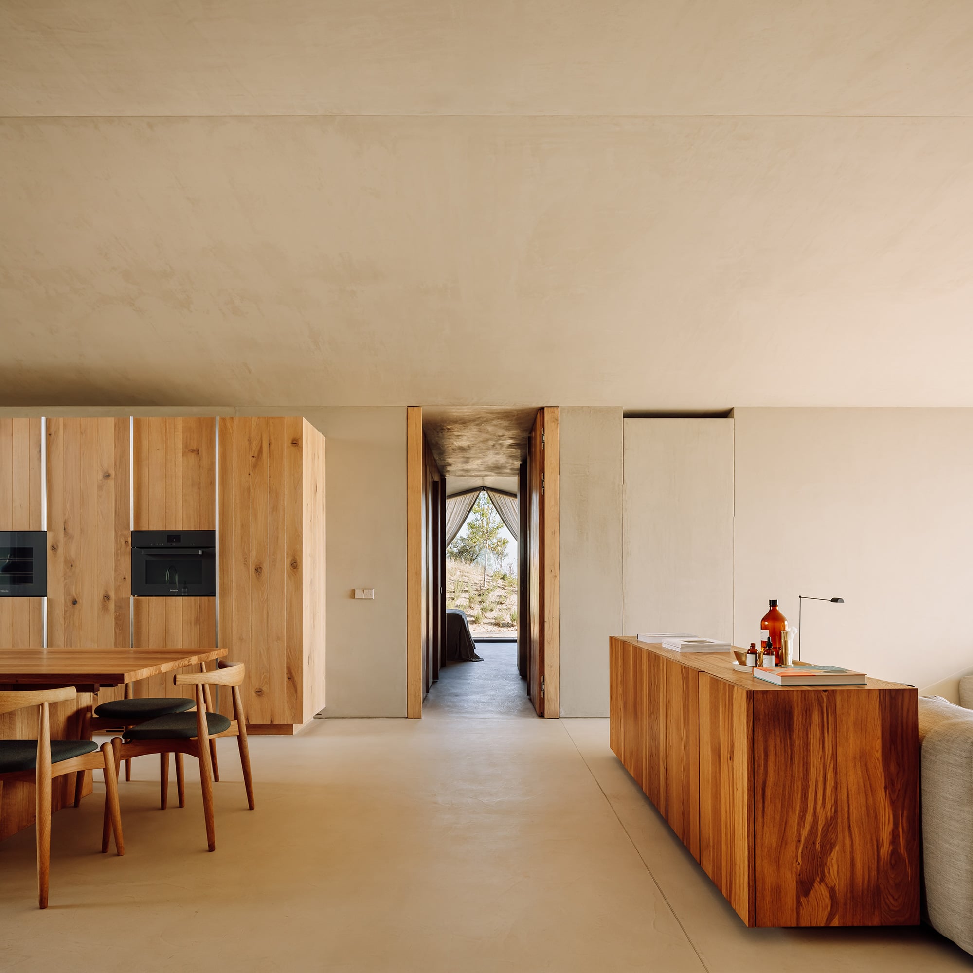 Pa.te.os, Europe — Minimalist kitchen and dining space with warm wood cabinetry, concrete flooring, and natural light through framed doorway opening to landscape.