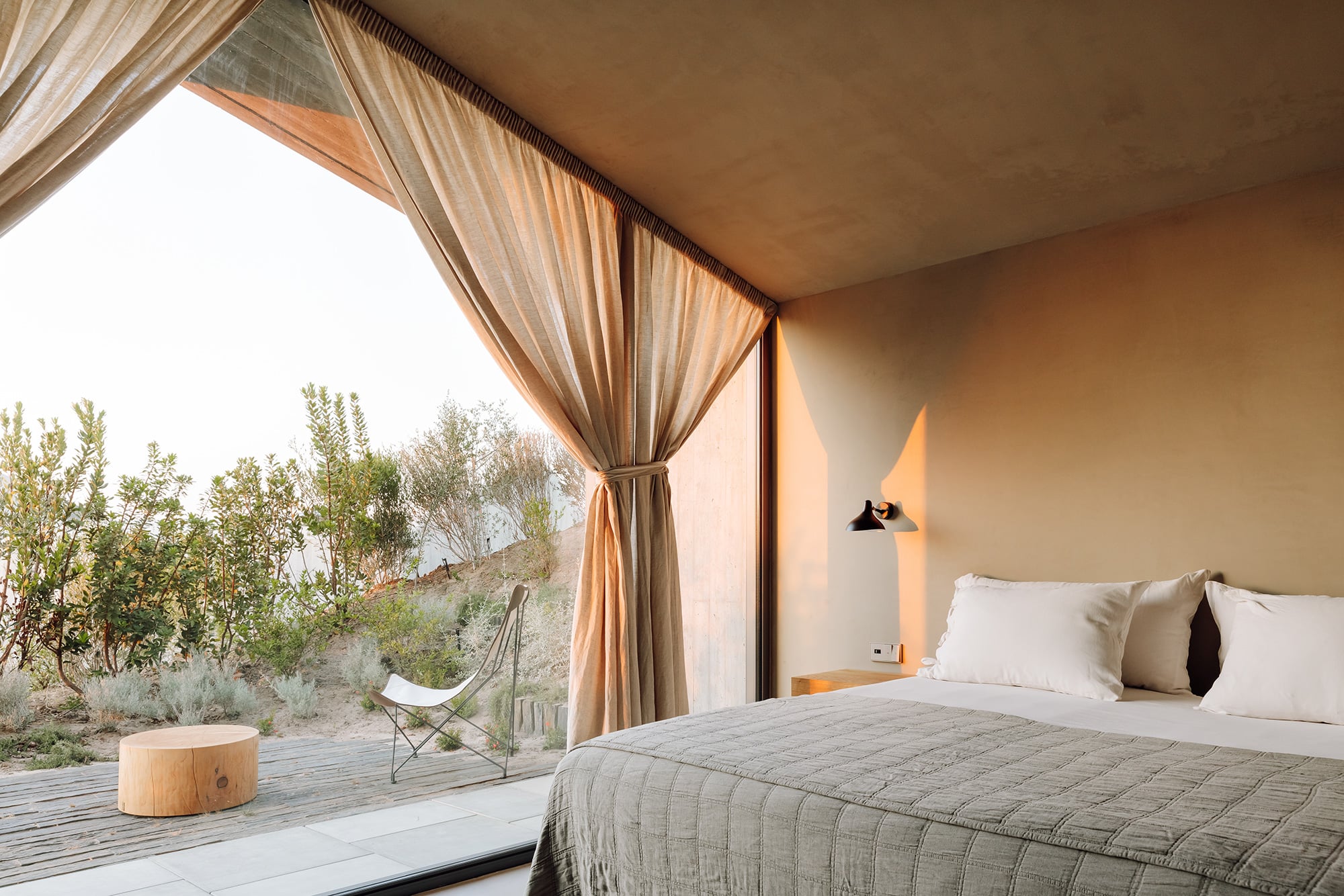 Pa.te.os, Europe — Minimalist bedroom with floor-to-ceiling glazing, natural light, linen bedding, warm earth tones, desert landscape view, modernist architecture.