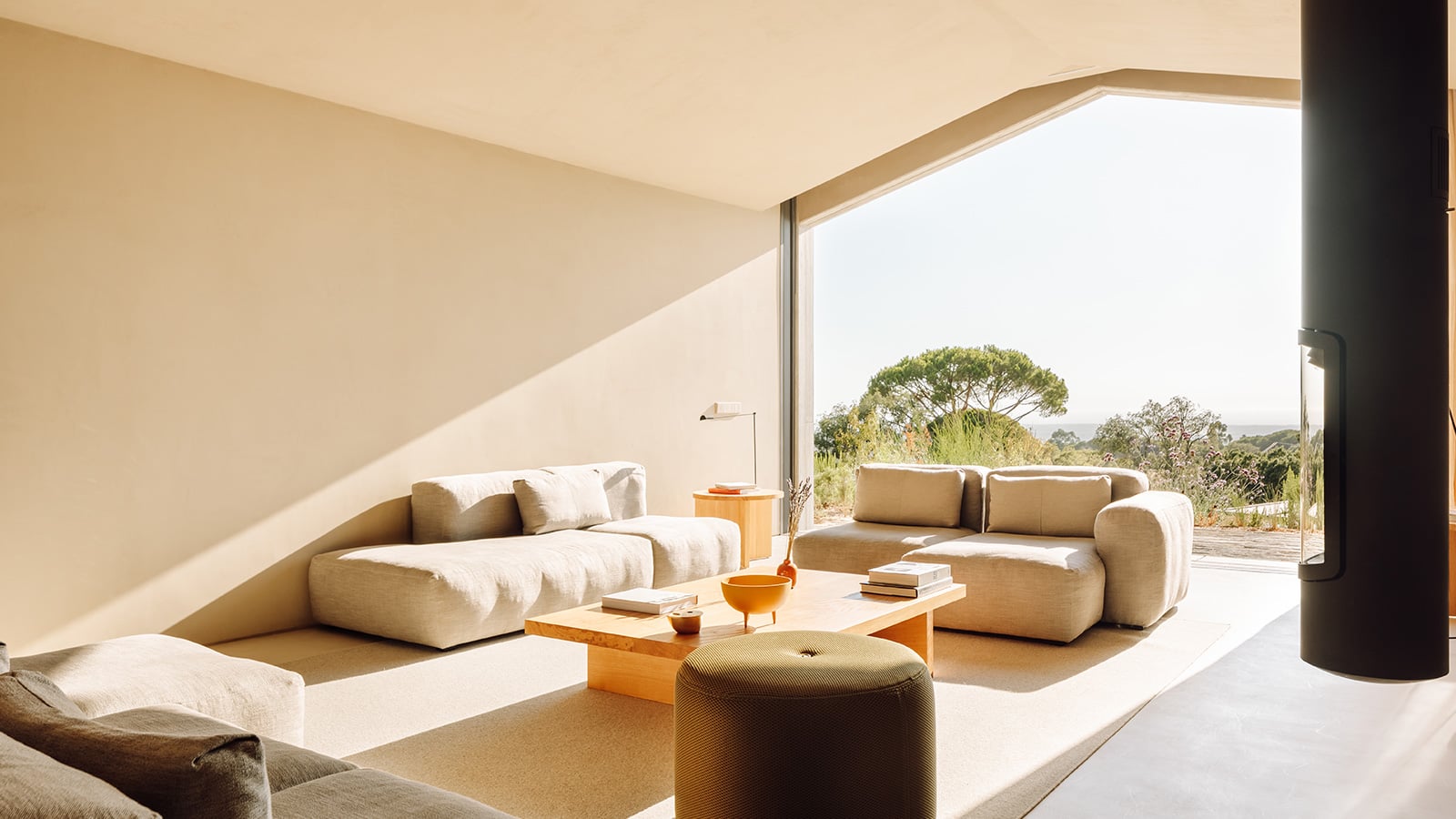 Pa.te.os, Europe — Minimalist villa living room with curved cream sofas, wooden coffee table, and floor-to-ceiling windows opening to Mediterranean landscape and terrace.