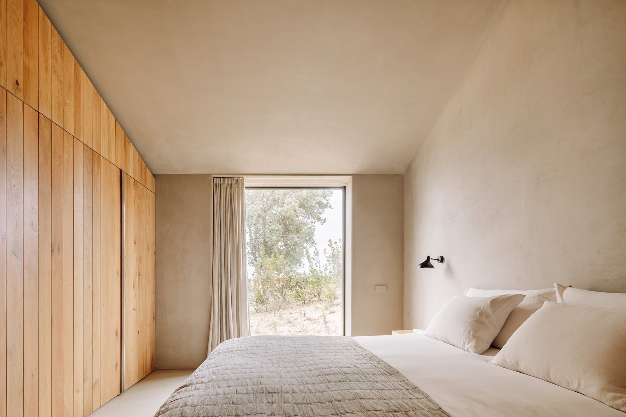 Pa.te.os, Europe — Minimalist bedroom with timber wardrobe, neutral plaster walls, linen bedding, and expansive glass doors opening to natural landscape light.