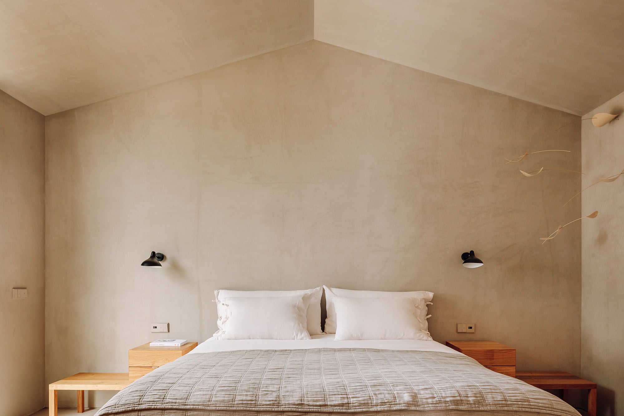 Pa.te.os, Europe — Minimalist bedroom with vaulted ceiling, natural plaster walls, wooden nightstands, white bedding, and sculptural black sconces.