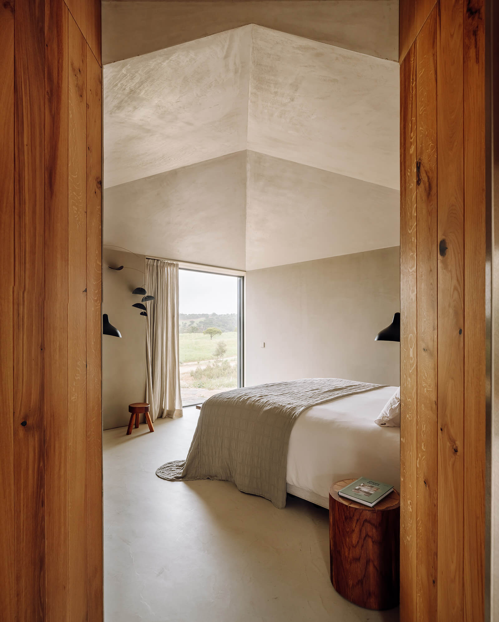 Pa.te.os, Europe — Minimalist bedroom with skylighted vaulted ceiling, timber-framed doors, platform bed, black pendant lights, and expansive countryside views through floor-to-ceiling glass.