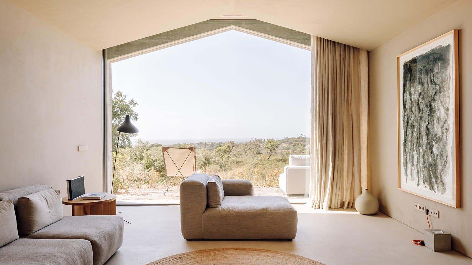 Pa.te.os, Europe — Minimalist living room with sculptural taupe seating, floor-to-ceiling windows framing rural landscape, warm natural light, polished concrete floors.