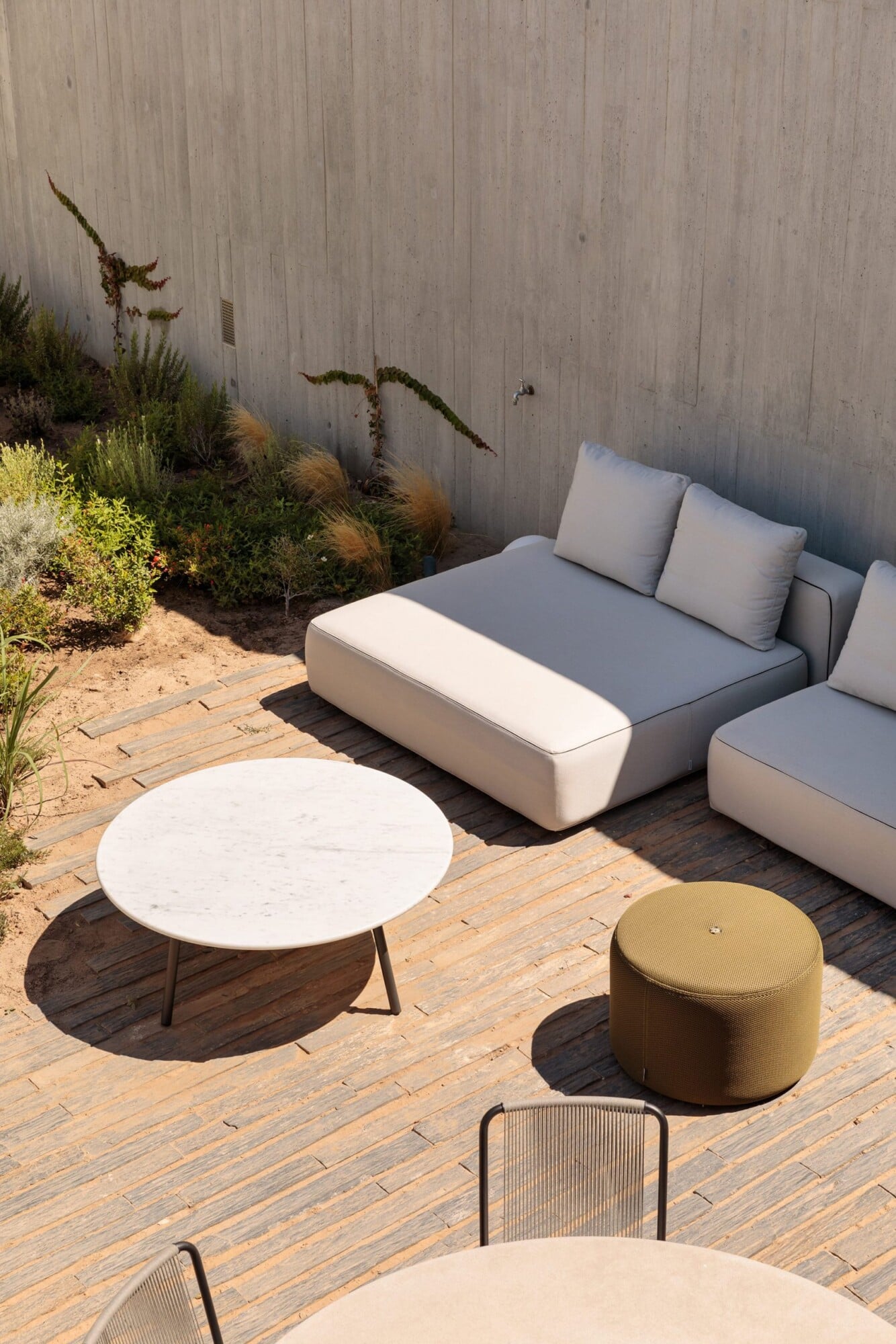Pa.te.os, Europe — Modern outdoor lounge with curved modular seating, marble side table, olive pouf, wooden deck, and desert plantings against concrete wall.
