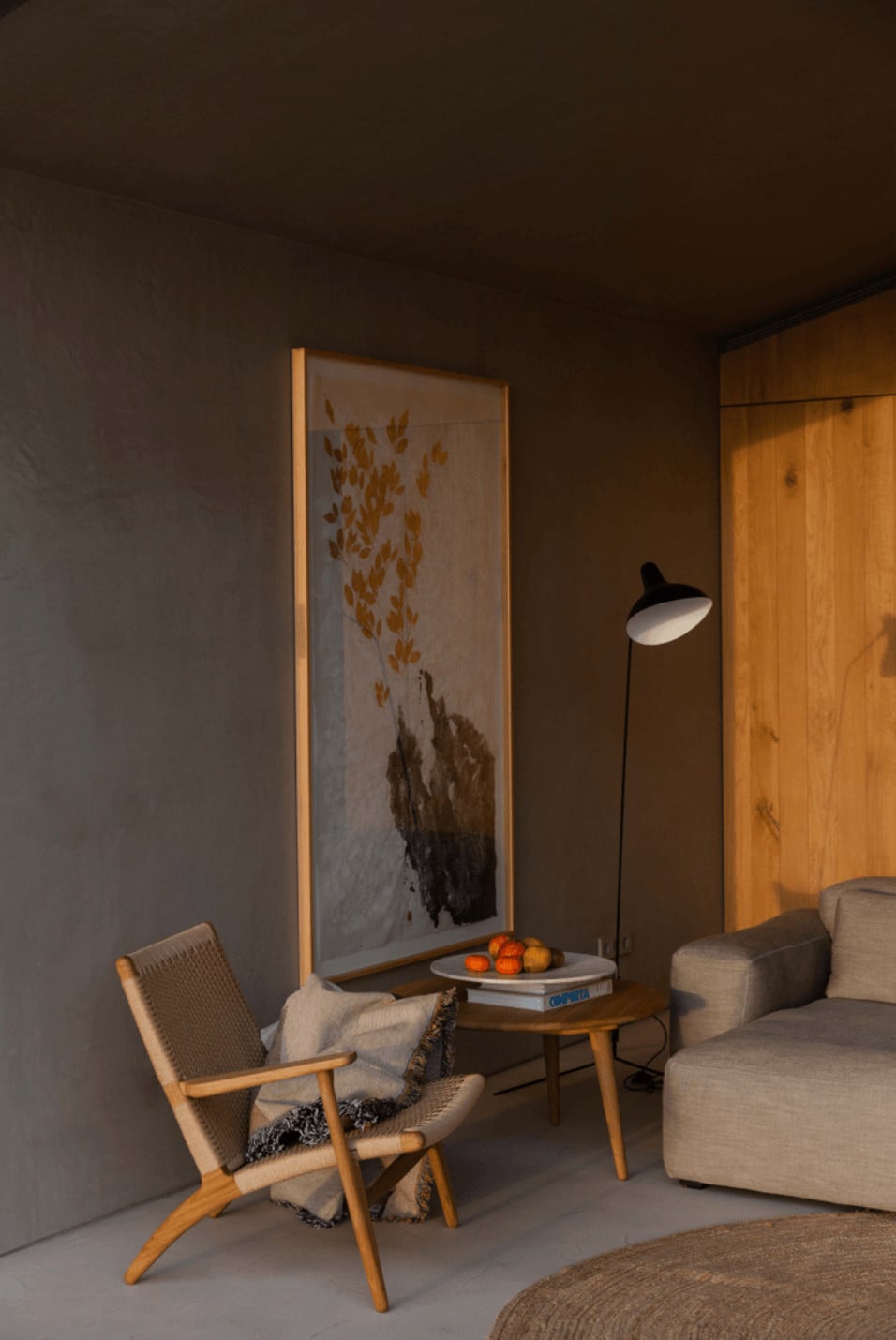 Pa.te.os, Europe — Minimalist hotel suite with wooden mid-century chair, linen sofa, framed botanical artwork, warm ambient lighting, and terracotta accents on side table.