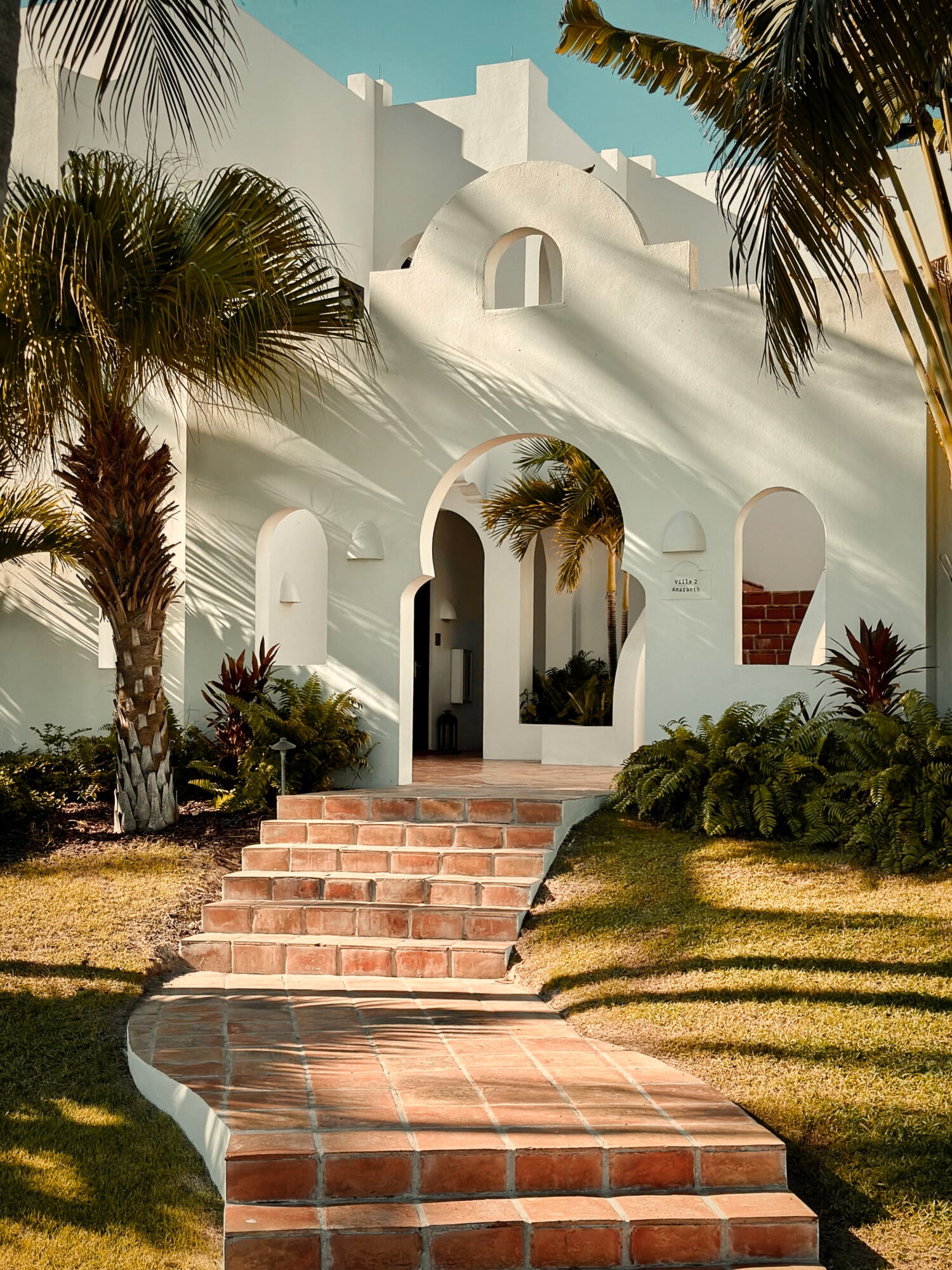 Cap Juluca, North America — White modernist villa with arched entrance, terracotta brick steps, palm trees, and dramatic shadow patterns under clear sky.