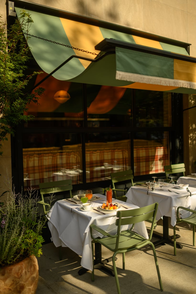 Nine Orchard, North America — Striped green and gold awning shades outdoor dining terrace with white-clothed tables, sage chairs, and floor-to-ceiling windows reflecting warm sunlight.