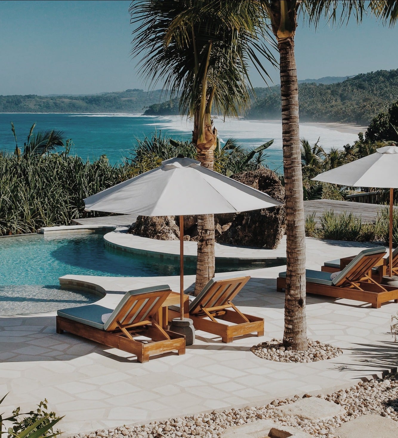 NIHI Sumba, Indonesia — Teak loungers with white umbrellas overlook turquoise infinity pool and pristine beach cove surrounded by tropical palms and lush hillside.
