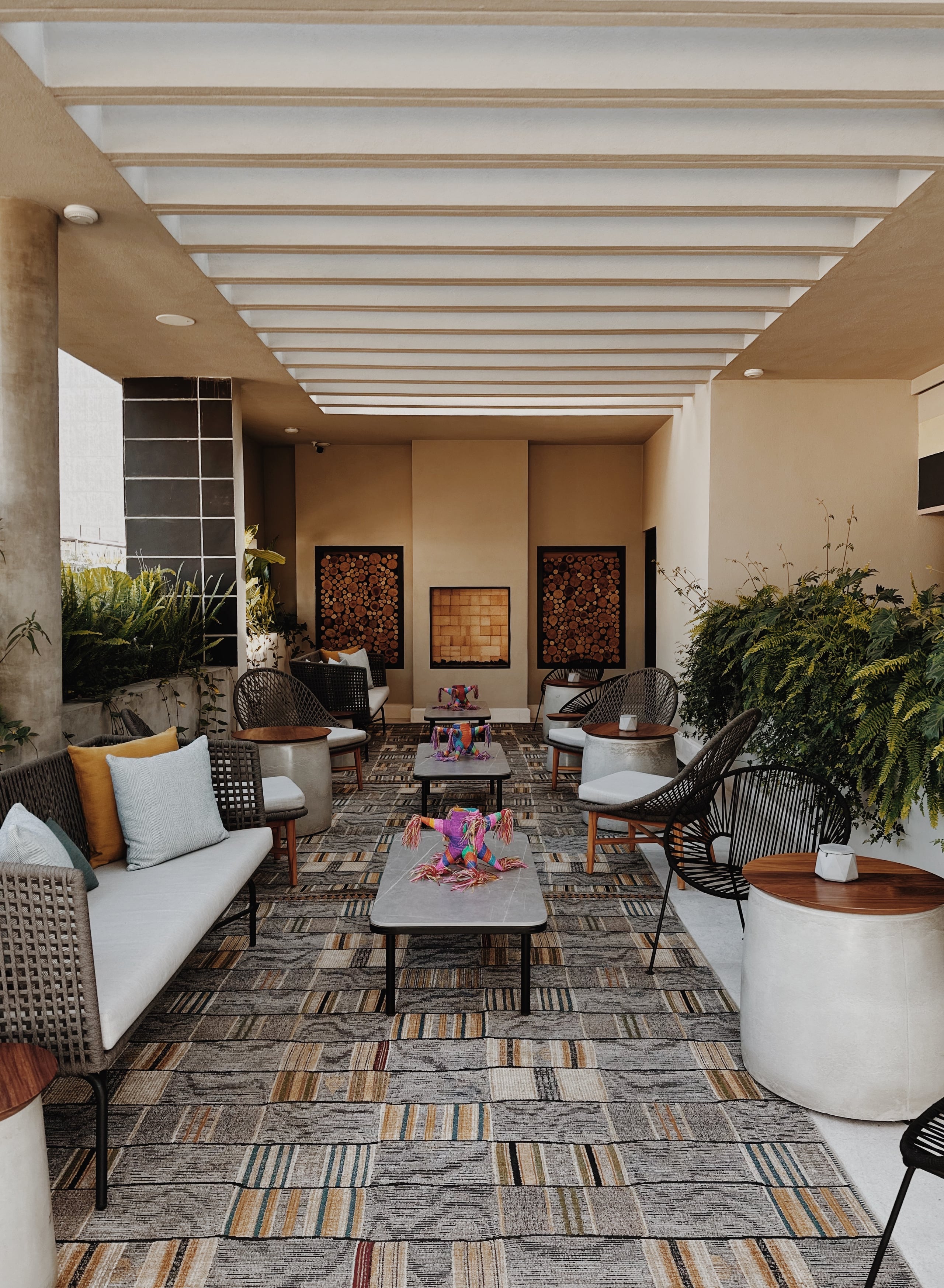 Campos Polanco, Mexico — Linear ceiling skylights illuminate a contemporary courtyard lounge with woven chairs, geometric rugs, potted greenery, and wood-relief wall art.