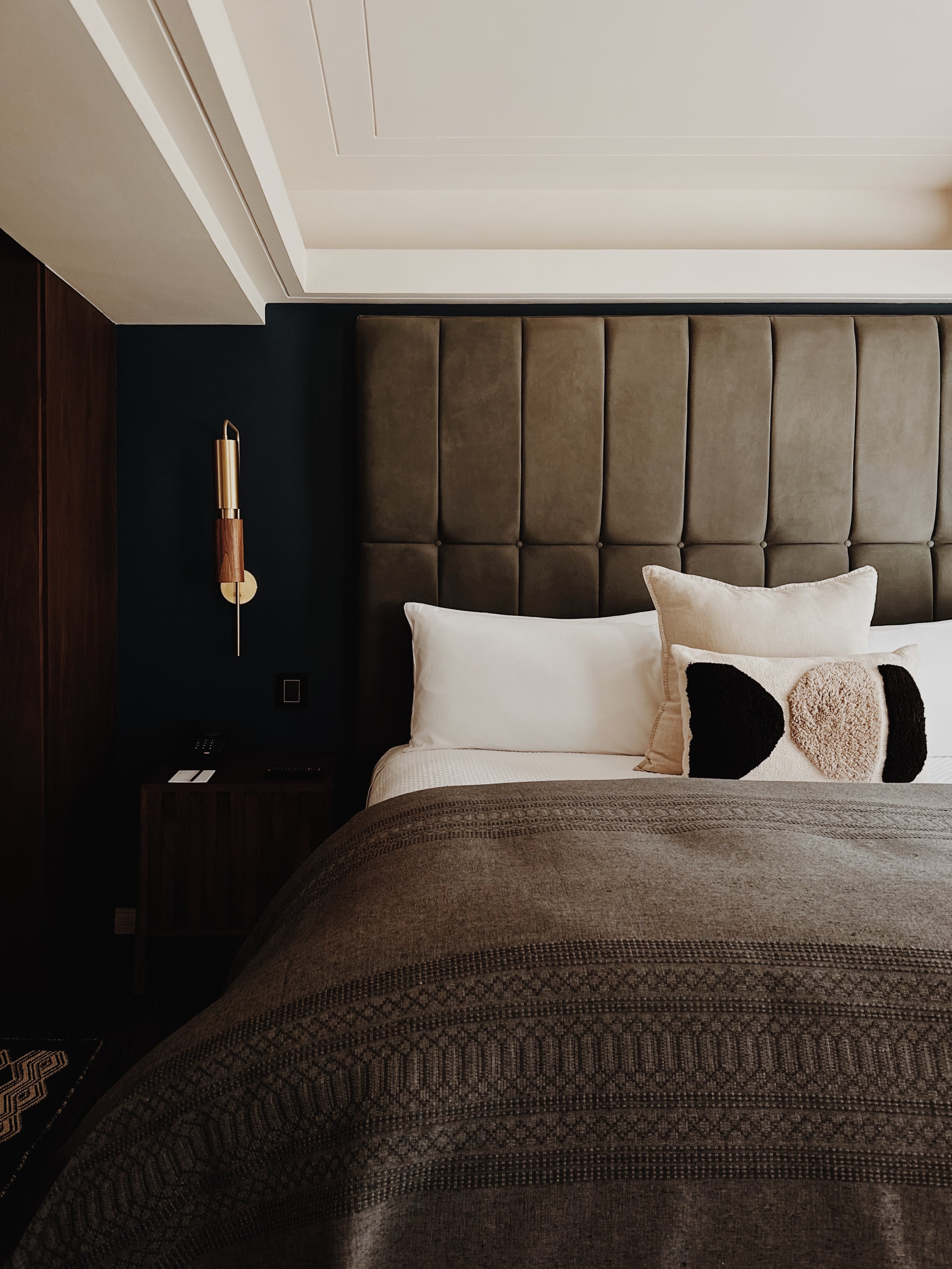 Campos Polanco, Mexico — Minimalist bedroom with taupe upholstered headboard, cream linens, geometric throw pillows, dark accent wall, brass sconce, coffered ceiling.