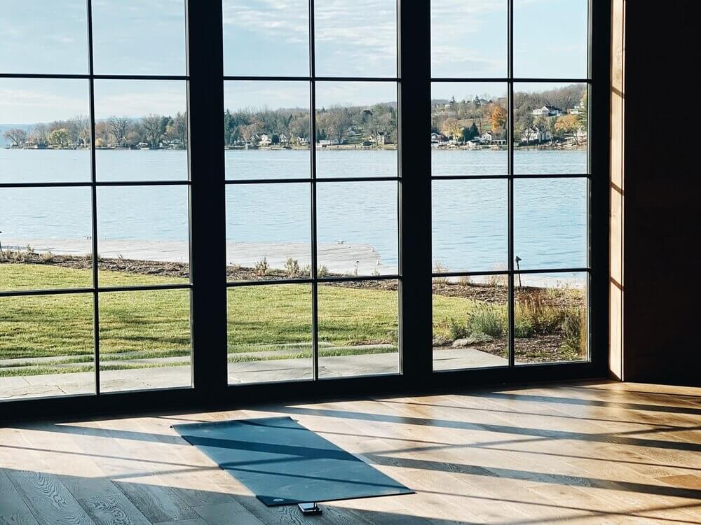 Willowbrook Spa, North America — Minimalist yoga studio with floor-to-ceiling windows, wooden flooring, blue mat, lakefront views, and residential shoreline landscape.