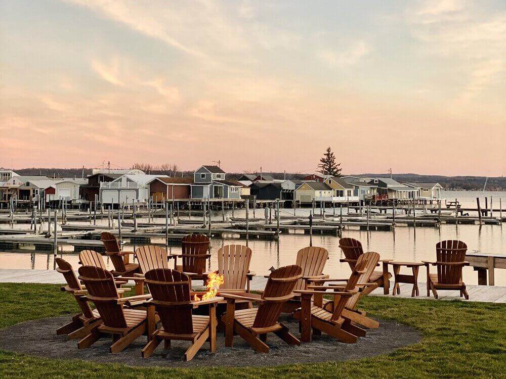 The Lake House, North America — Adirondack chairs arranged around fire pit on lawn overlooking waterfront boathouses at dusk, natural wood furniture with calm reflective water and residential architecture beyond.