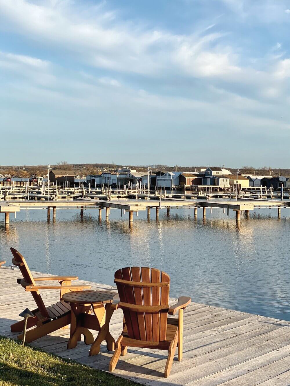 The Lake House, North America — Wooden Adirondack chairs on waterfront deck overlooking calm bay with historic boathouses and pier reflections under clear sky.