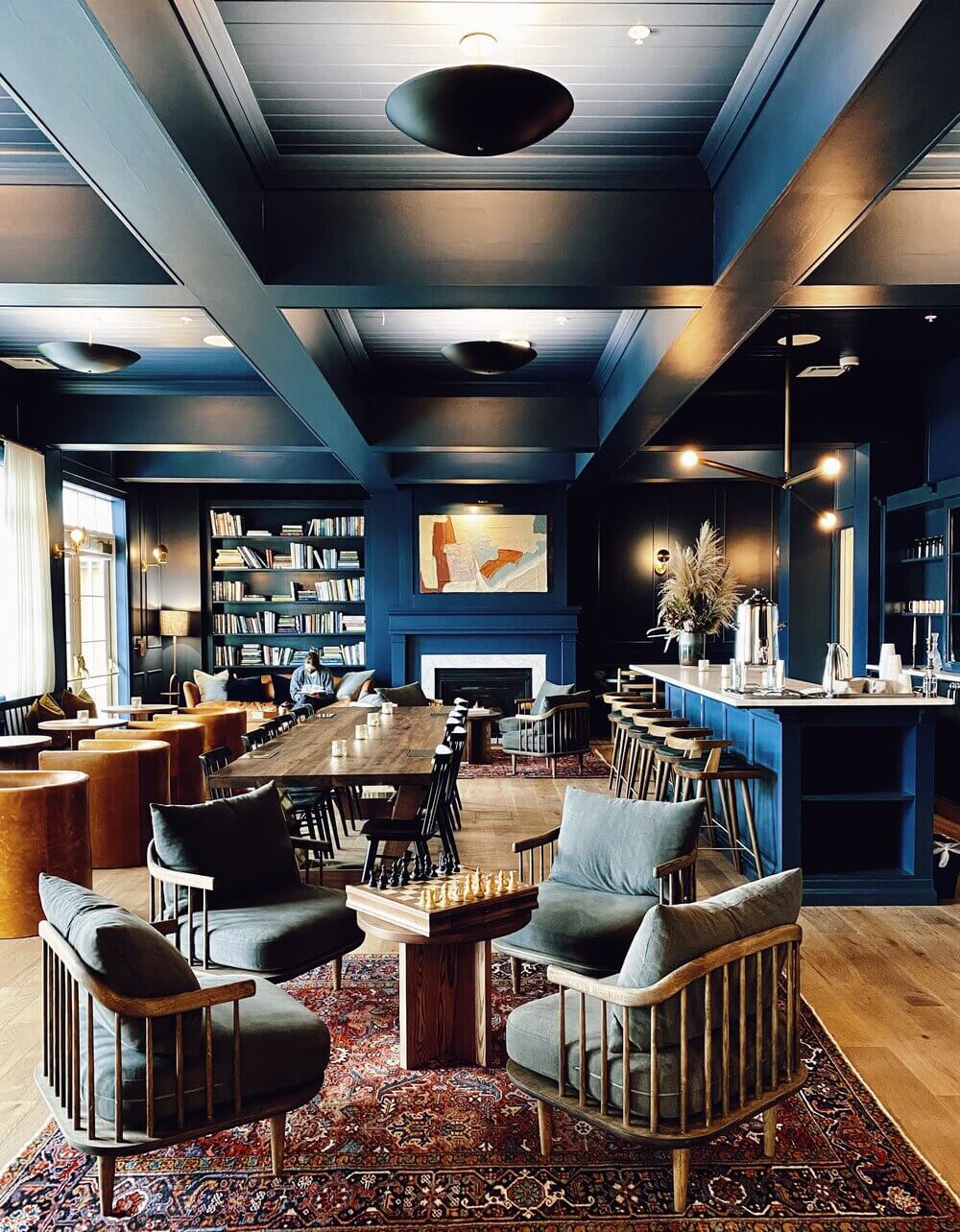 The Lake House — Lake House, North America — Deep blue library lounge with coffered ceiling, wood-framed chairs, fireplace, and chess table under warm pendant lighting.