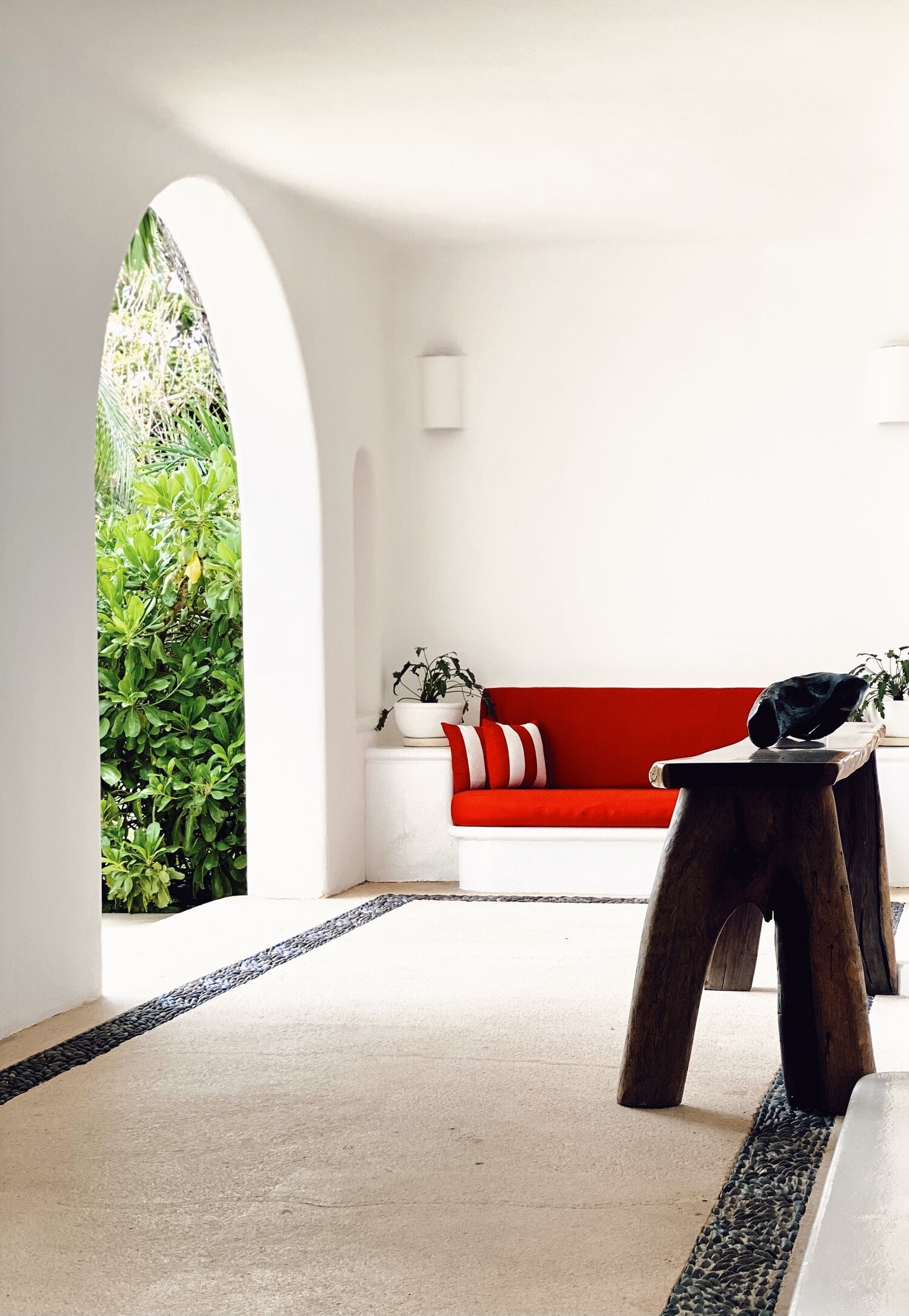Hotel Esencia, Mexico — Minimalist interior with arched window framing lush greenery, red sofa with striped pillow, dark wooden table, and black pebble floor detail.
