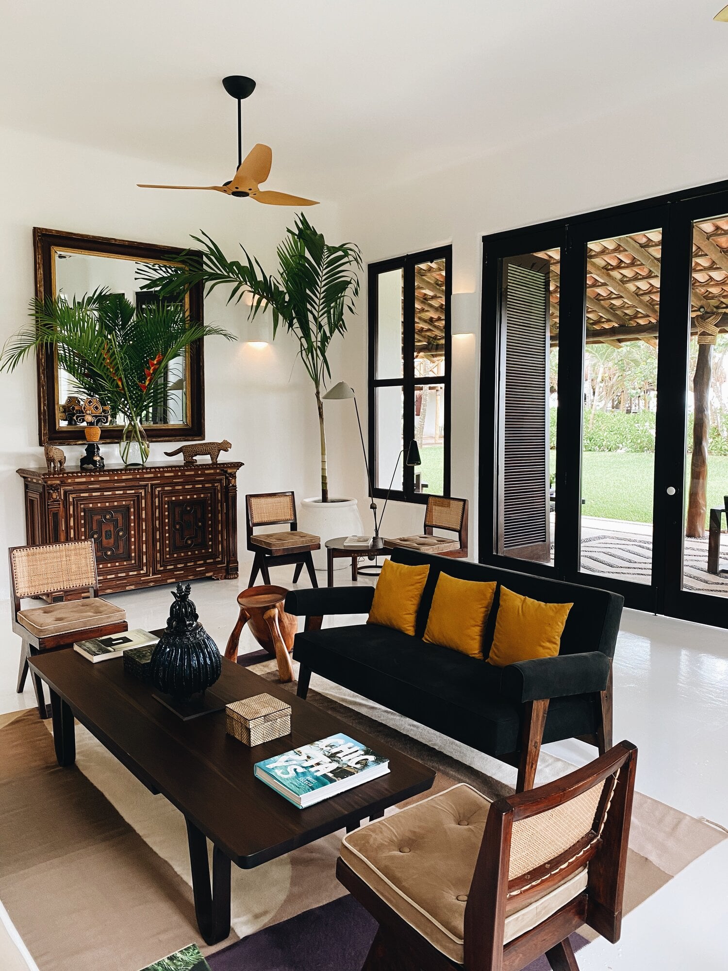 Hotel Esencia, Mexico — Mid-century modern living room with black sofa, wooden furniture, golden cushions, potted palms, and black-framed doors opening to tropical gardens.