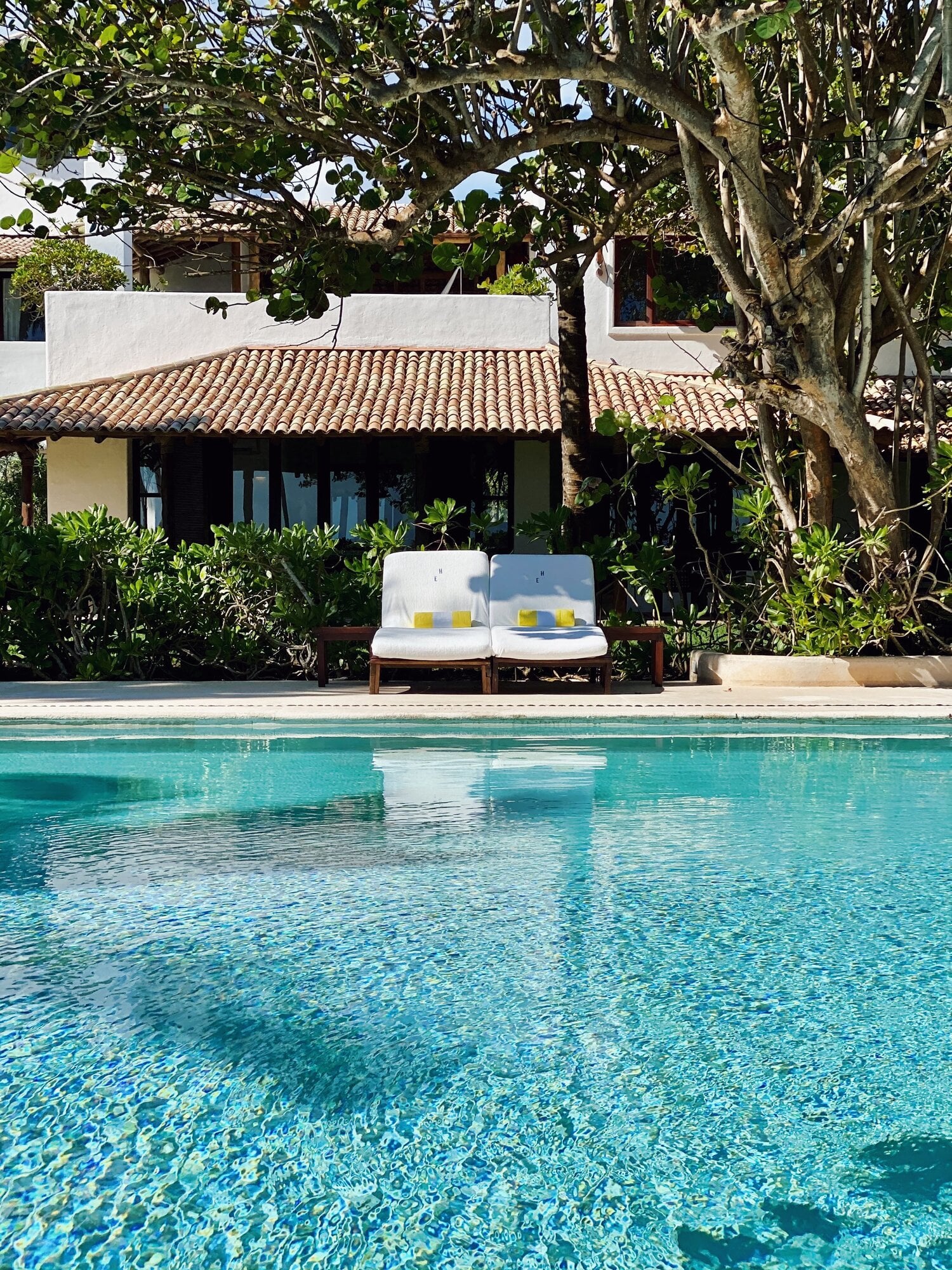 Hotel Esencia, Mexico — Turquoise pool with limestone terrace, colonial-style casita with clay tile roof, mature tree canopy, daybed seating.