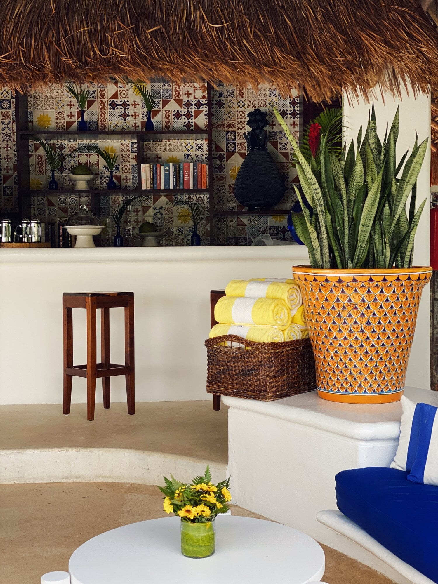 Hotel Esencia, Mexico — Bohemian-modern beach lounge with patterned tile backdrop, woven ceiling, potted succulents, yellow rolled towels, and blue cushioned seating.