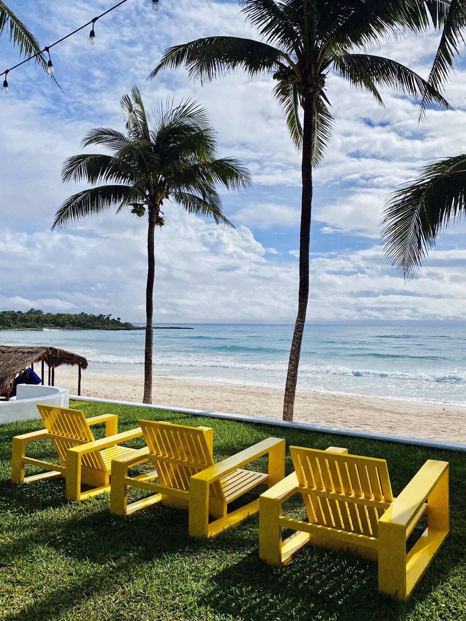 Hotel Esencia, Mexico — Bright yellow Adirondack chairs on manicured lawn facing turquoise ocean, palm trees, white sand beach, and clouded sky.