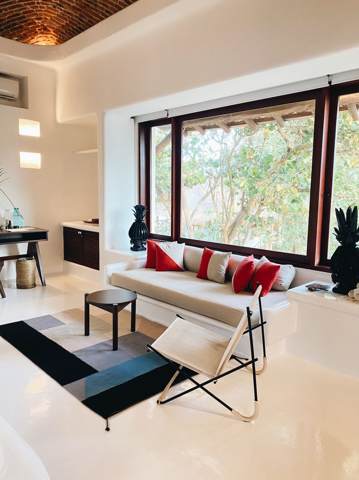 Hotel Esencia, Mexico — Modernist living room with cream sectional, red velvet cushions, dark wood-framed windows overlooking trees, geometric area rug, sculptural lighting, and warm ambient glow.