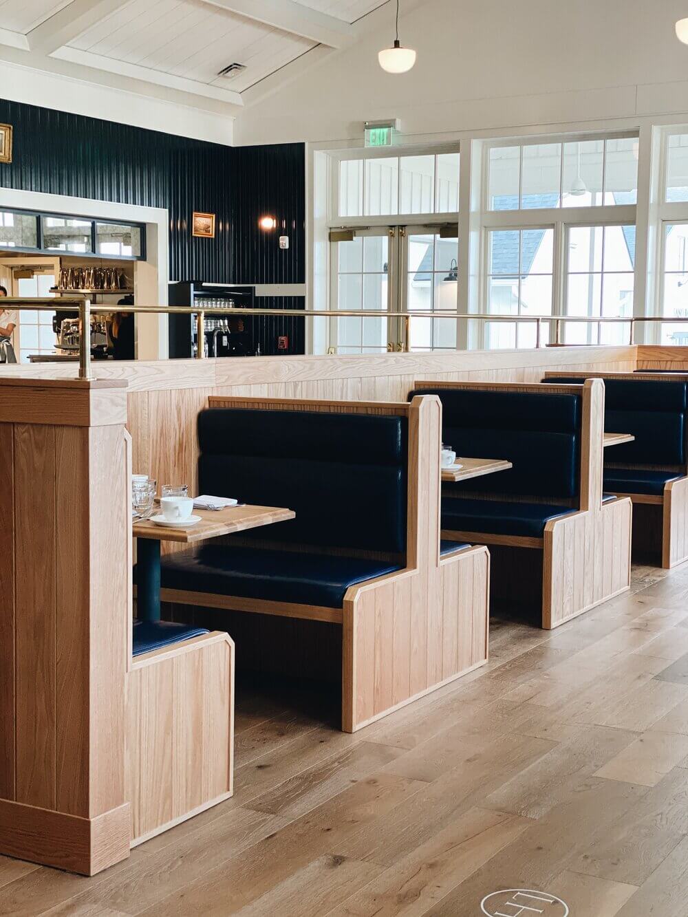 The Lake House, North America — Light wood booth seating with navy leather, clerestory windows overlooking water, modern coastal restaurant interior with black corrugated accent wall.