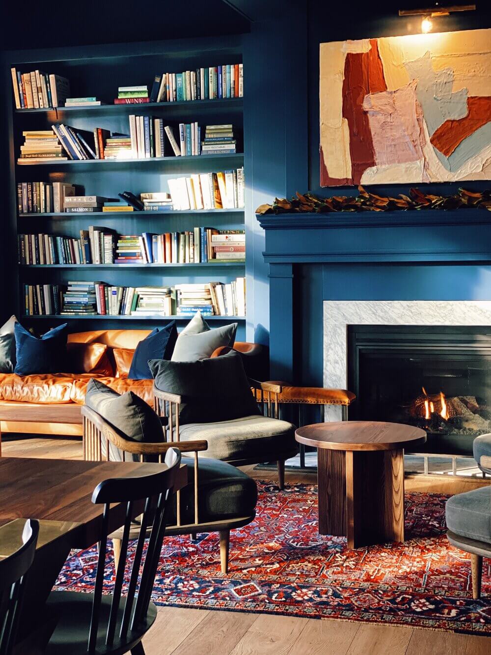 The Lake House, North America — Deep blue library with built-in shelving, fireplace, mid-century furniture, warm wood accents, and patterned rug creating intimate reading lounge.