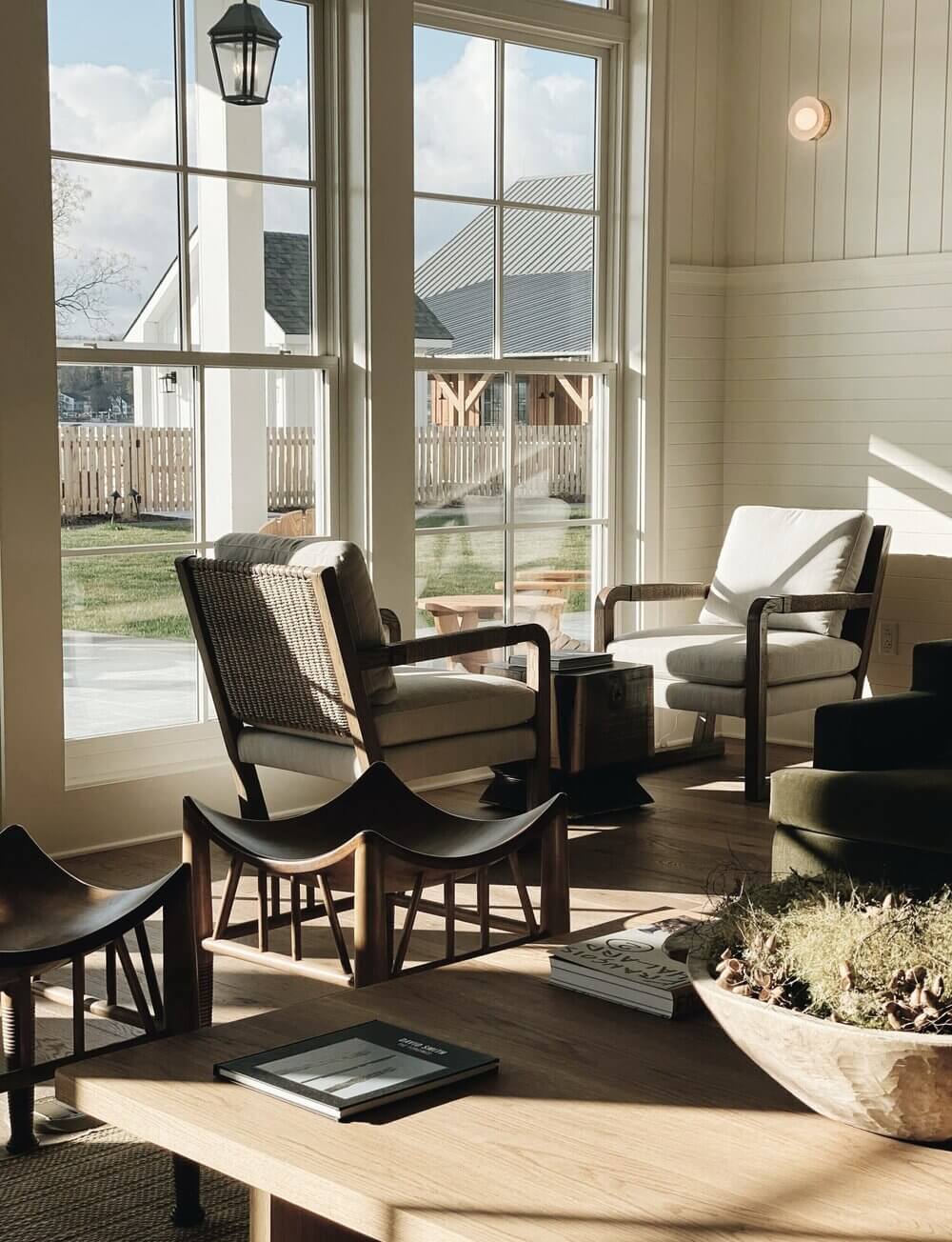 The Lake House, North America — Modern sunlit lounge with rattan and black wood chairs, white pillows, herringbone ceiling, floor-to-ceiling windows overlooking manicured grounds and distant architecture.