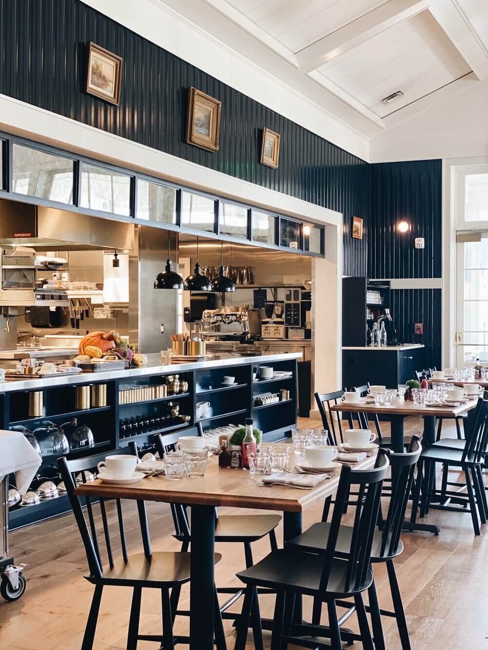 Rose Tavern, North America — Dark-paneled café with open kitchen, wooden tables, black Windsor chairs, skylit ceiling, brass pendant lights, and vintage framed artwork.