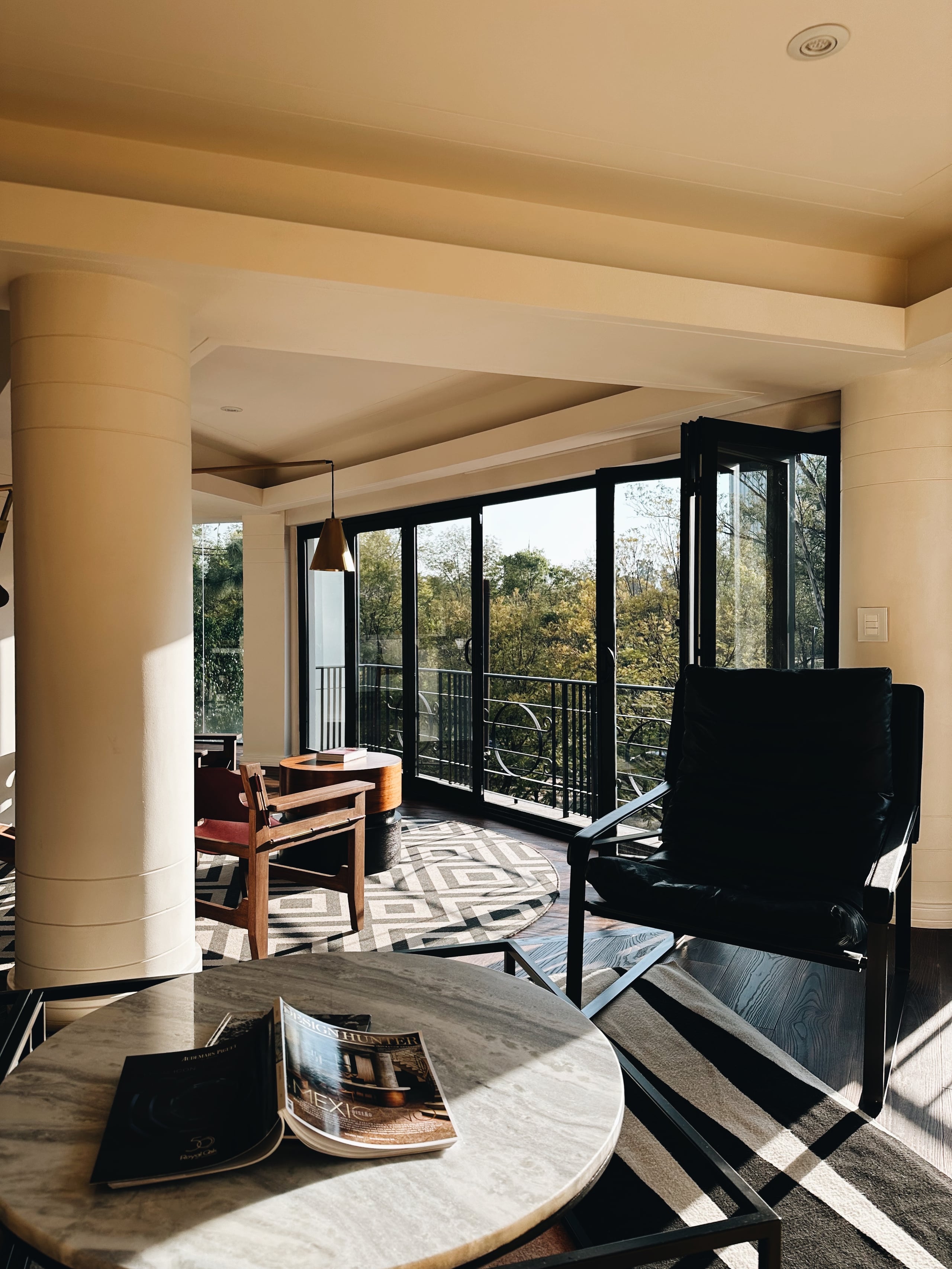 Campos Polanco, Mexico — Modern living space with floor-to-ceiling black-framed glass doors, natural light, geometric patterned rug, wood furnishings, and forest views from elevated terrace.