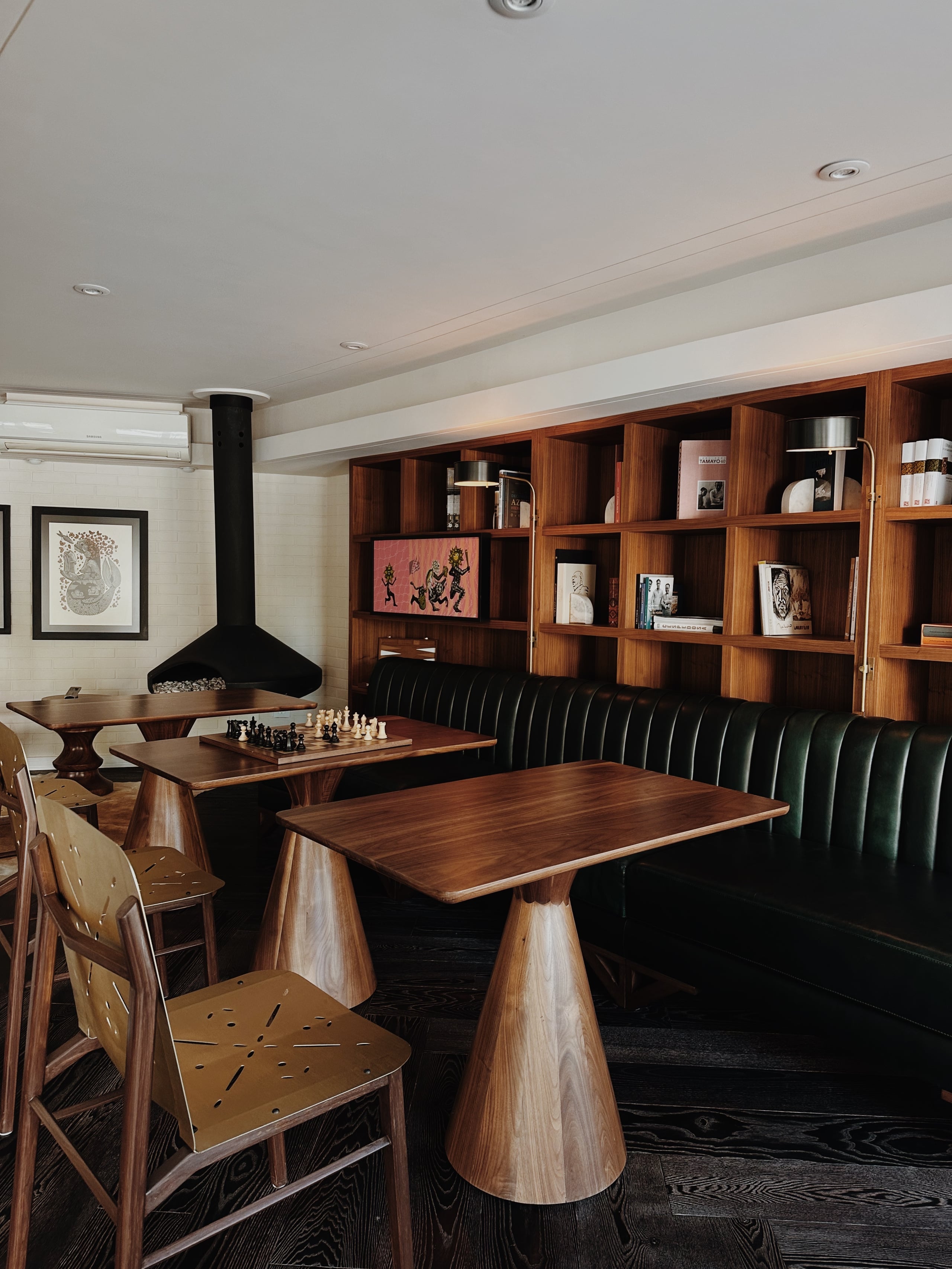 Campos Polanco, Mexico — Mid-century wood lounge with green leather banquette, walnut tables, chess set, black fireplace, and built-in shelving displaying art and objects.