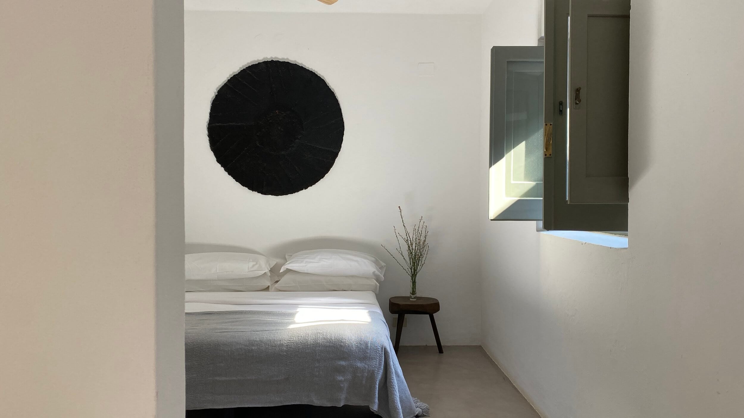 Parco Dei Sesi, Europe — Minimalist bedroom with white walls, black circular artwork, linen bedding, wooden side table with dried grasses, and open window flooding natural light.