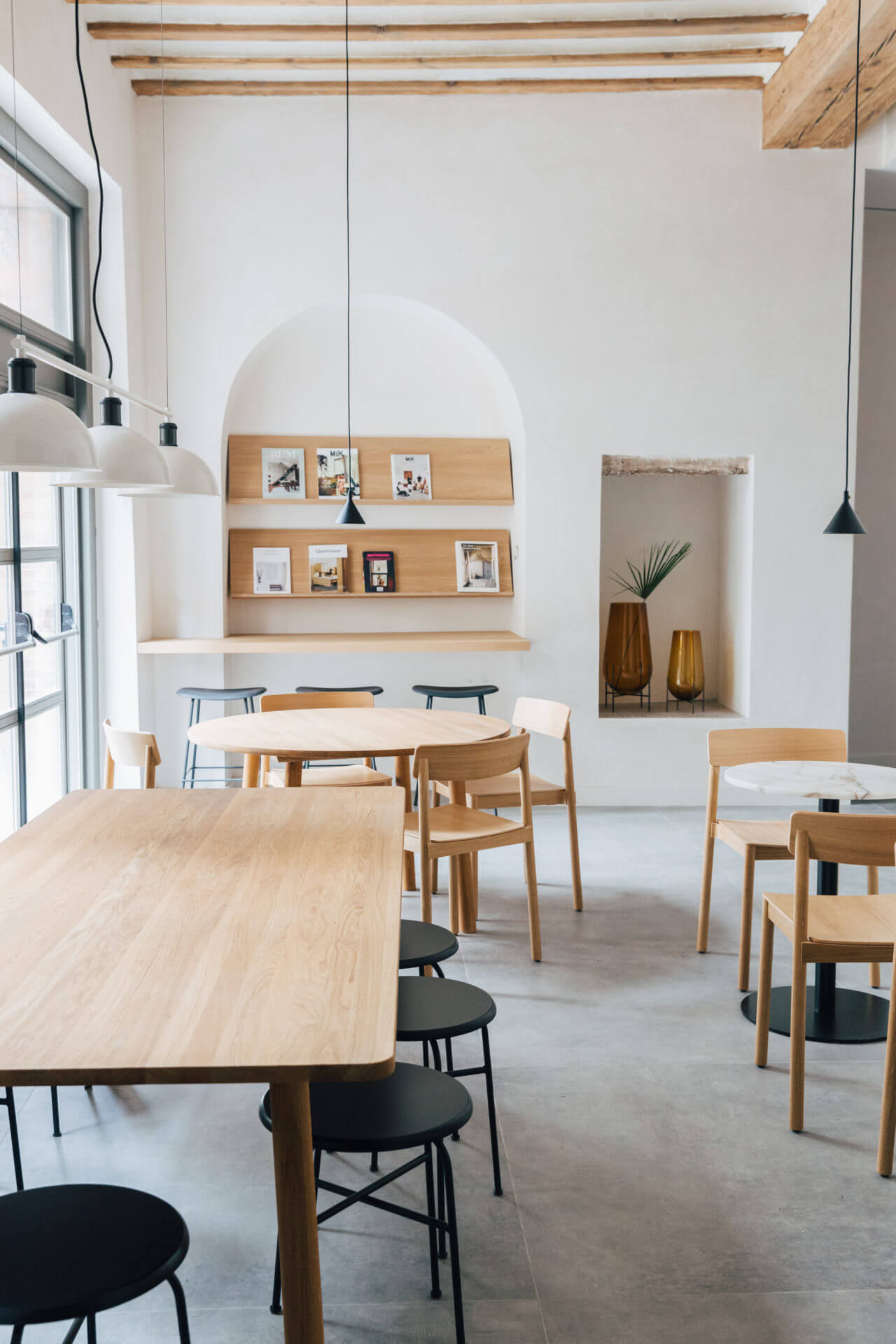 Plácido y Grata, Europe — Minimalist dining space with light oak tables, black chairs, exposed beams, and built-in wooden shelving displaying curated objects beneath arched alcove.