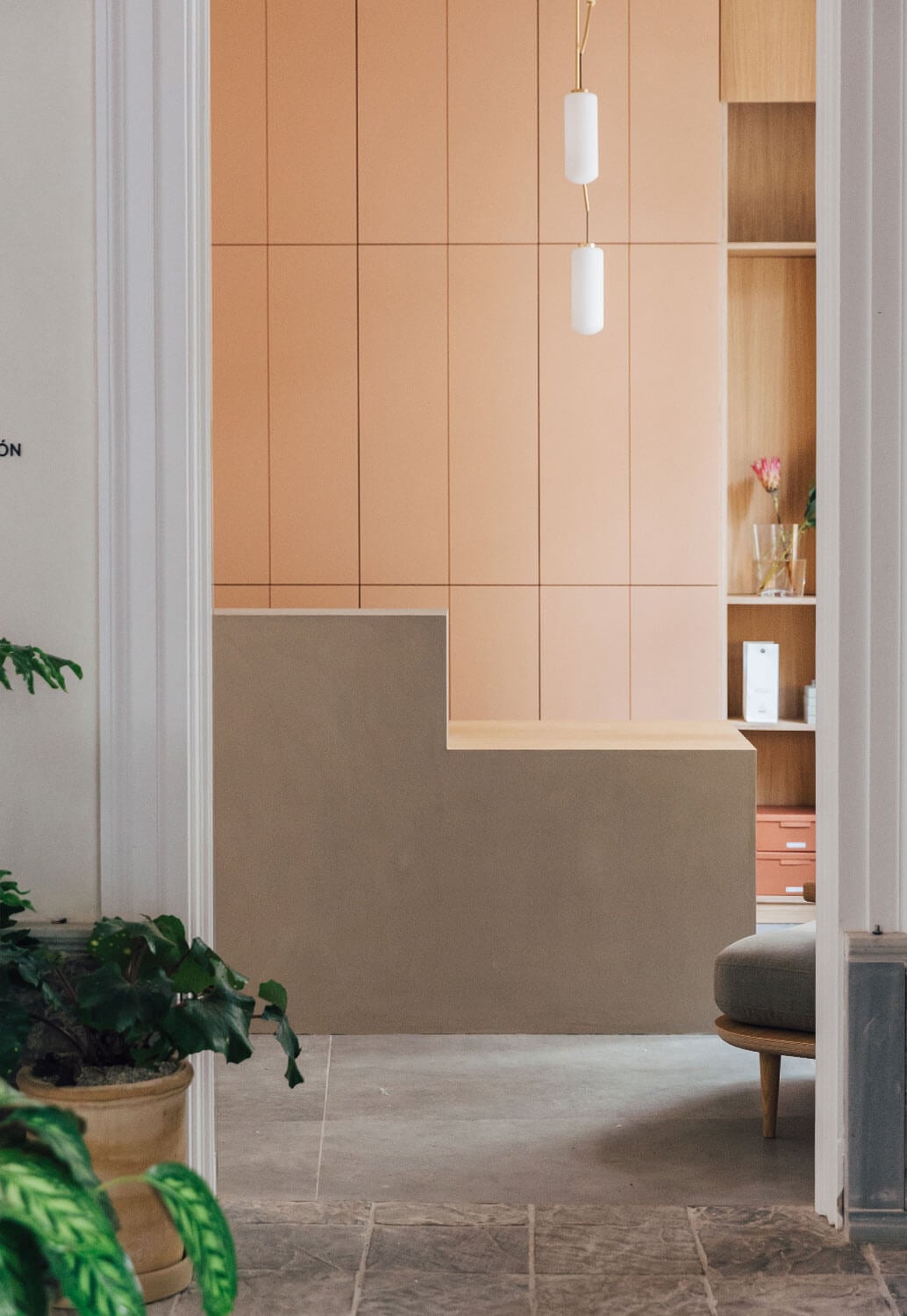 Plácido y Grata, Europe — Minimalist reception desk with peach-toned paneling, pendant lights, wooden shelving, and potted plants in warm-lit hospitality space.