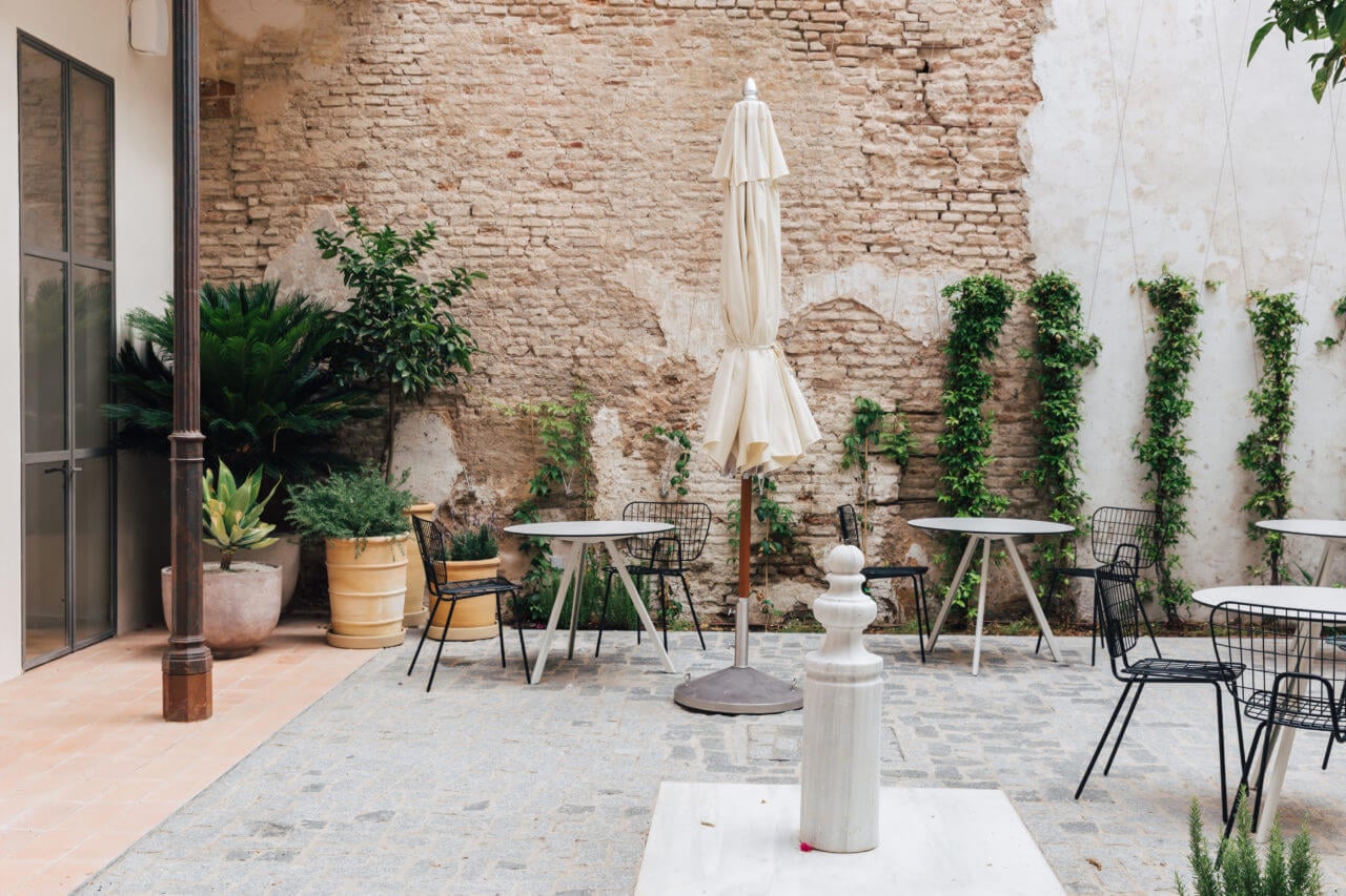 Plácido y Grata, Europe — Courtyard with cream parasol, black modern chairs, terracotta and stone flooring, climbing ivy on exposed brick walls, Mediterranean hotel design.