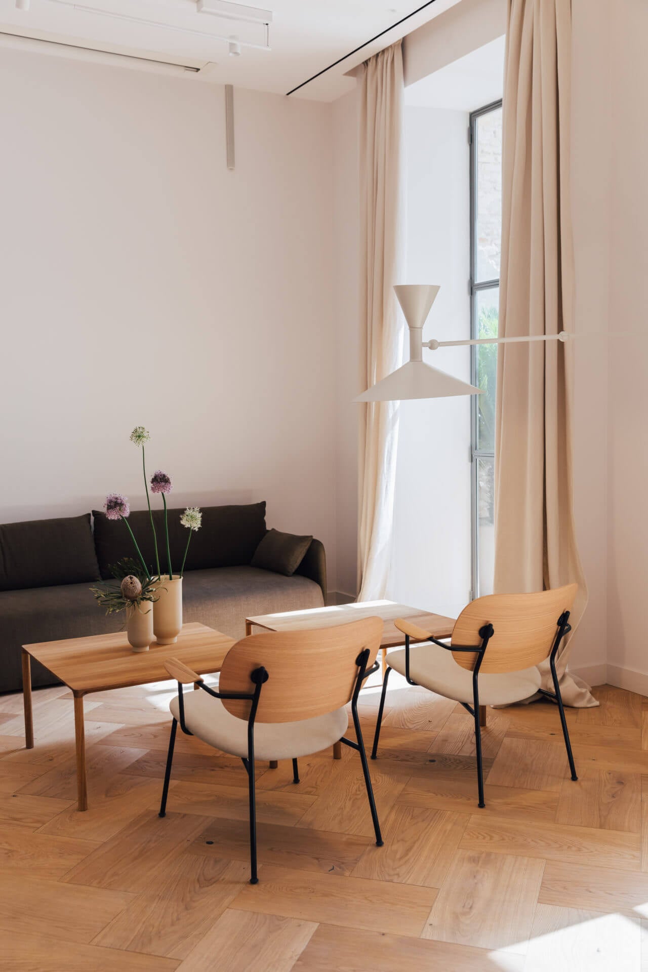 Plácido y Grata, Europe — Minimalist lounge with natural wood chairs, black metal frames, cream upholstery, beige sofa, wooden table with flowers, floor-to-ceiling windows, warm neutral palette.