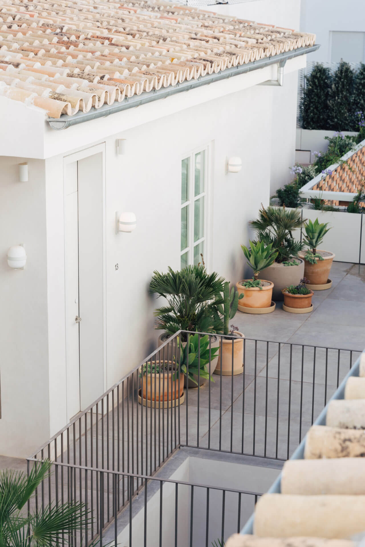 Plácido y Grata, Europe — Minimalist courtyard with terra-cotta planters, white-walled architecture, black metal railings, and terracotta tile roof detail.