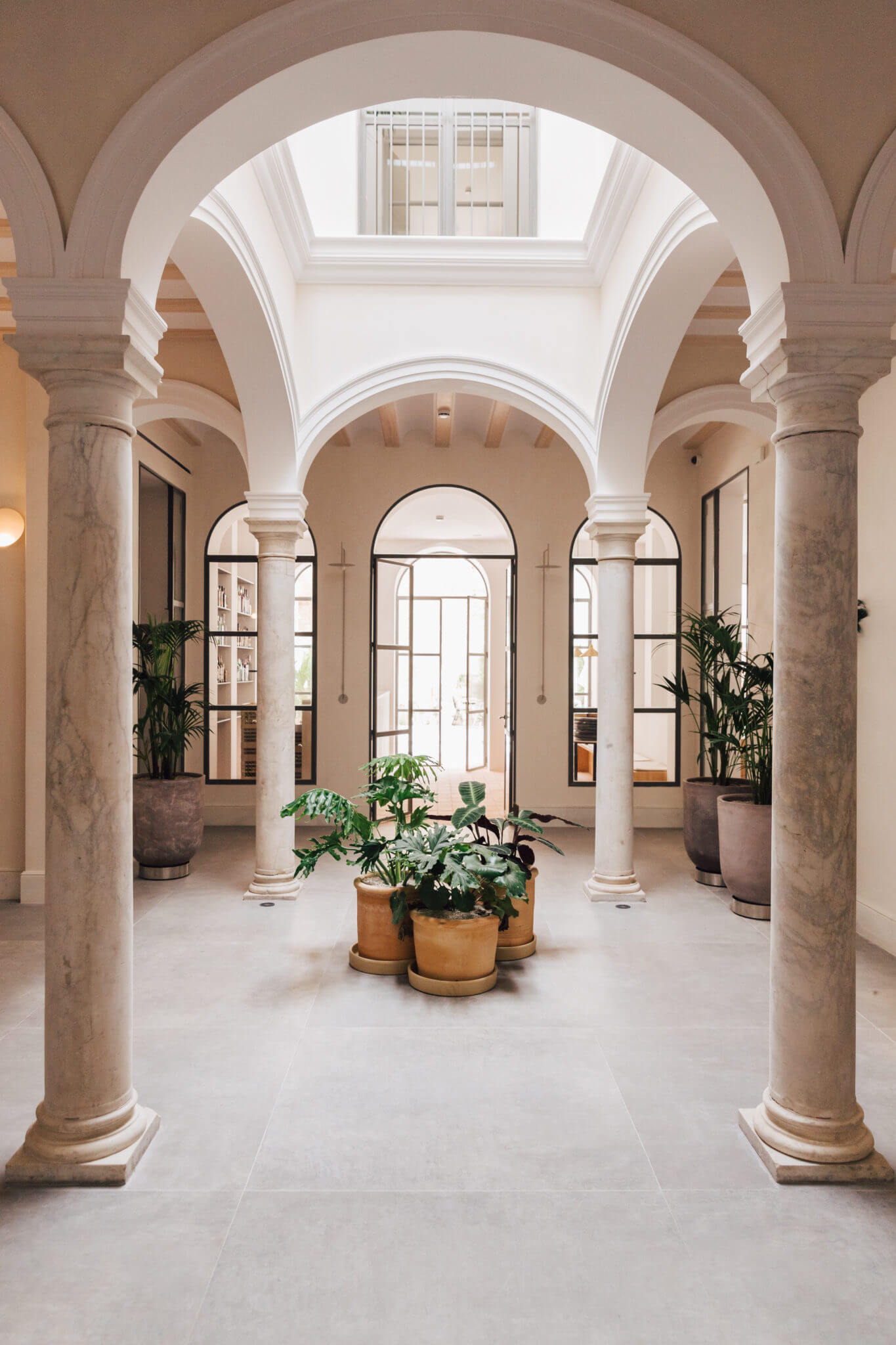 Plácido y Grata, Europe — Neoclassical courtyard with travertine columns, arched vaults, terracotta planters, and gridded doors flooding natural light through skylights.