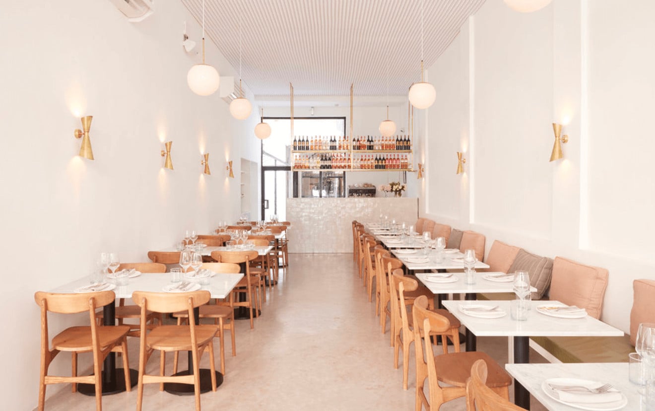 plus61, Africa — Minimalist dining room with natural wood chairs, white marble tables, brass pendant lights, and floor-to-ceiling wine display behind stone bar counter.