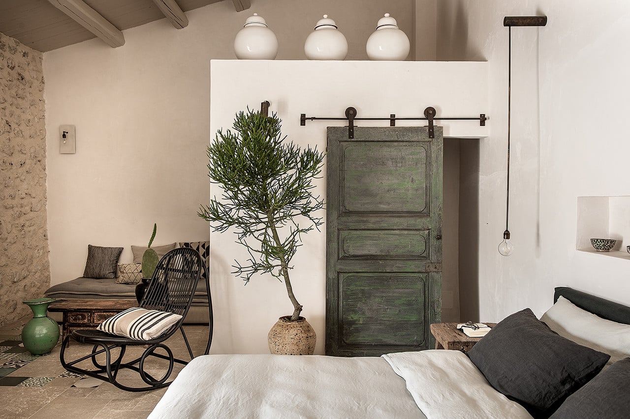 Dimora delle Balze, Europe — Minimalist bedroom with terracotta walls, barn door, bonsai plant, rocking chair, pendant light, and natural stone flooring.