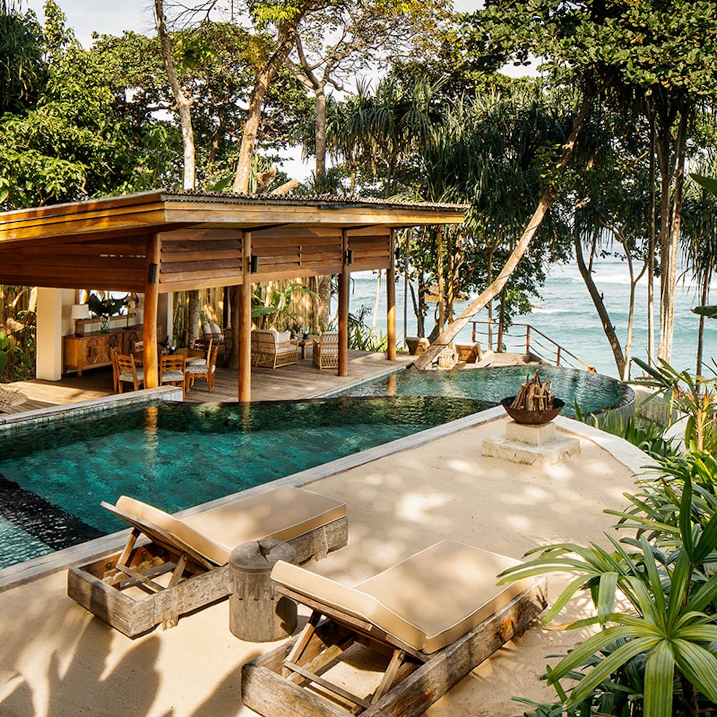 NIHI Sumba, Asia — Teak pavilion with infinity pool overlooking turquoise ocean, weathered lounge chairs on sand, tropical trees, minimalist coastal architecture.