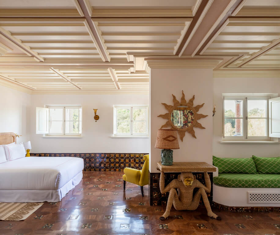 Vermelho, Europe — Sunlit bedroom with coffered ceiling, colorful tile accents, woven sun mirror, green sofa, and sculptural wooden monkey figure.