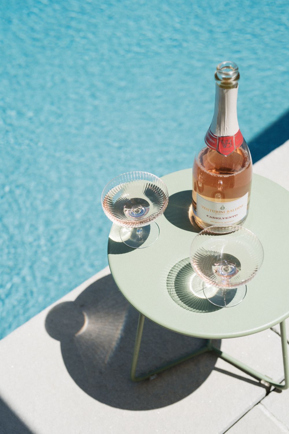 Pool Bar, Europe — Rosé champagne bottle and two coupe glasses on white table beside turquoise swimming pool in bright sunlight.