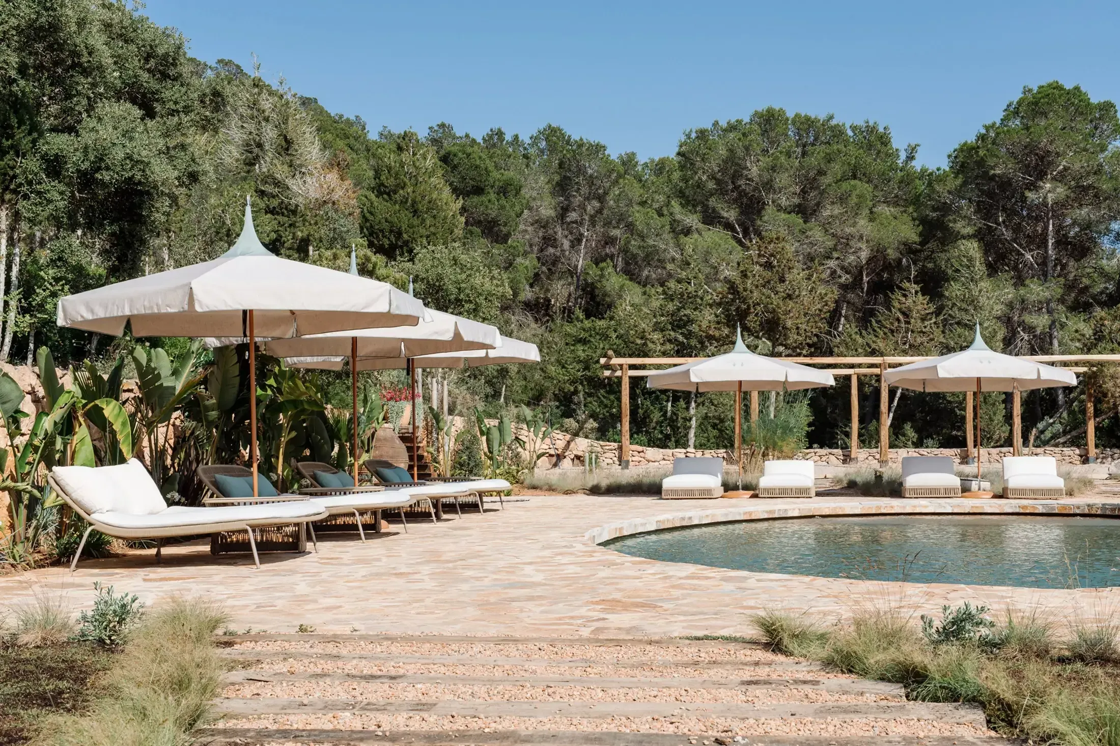 Finca La Inika, Balearic Islands — Poolside lounge area with canvas umbrellas, wooden sunbeds, and Mediterranean pine forest backdrop.