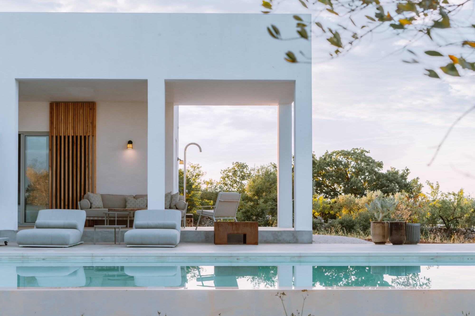 Villa Quercia, Italy — Minimalist poolside terrace with white architecture, wooden slat screen, pale stone loungers, and Mediterranean landscape views at dusk.