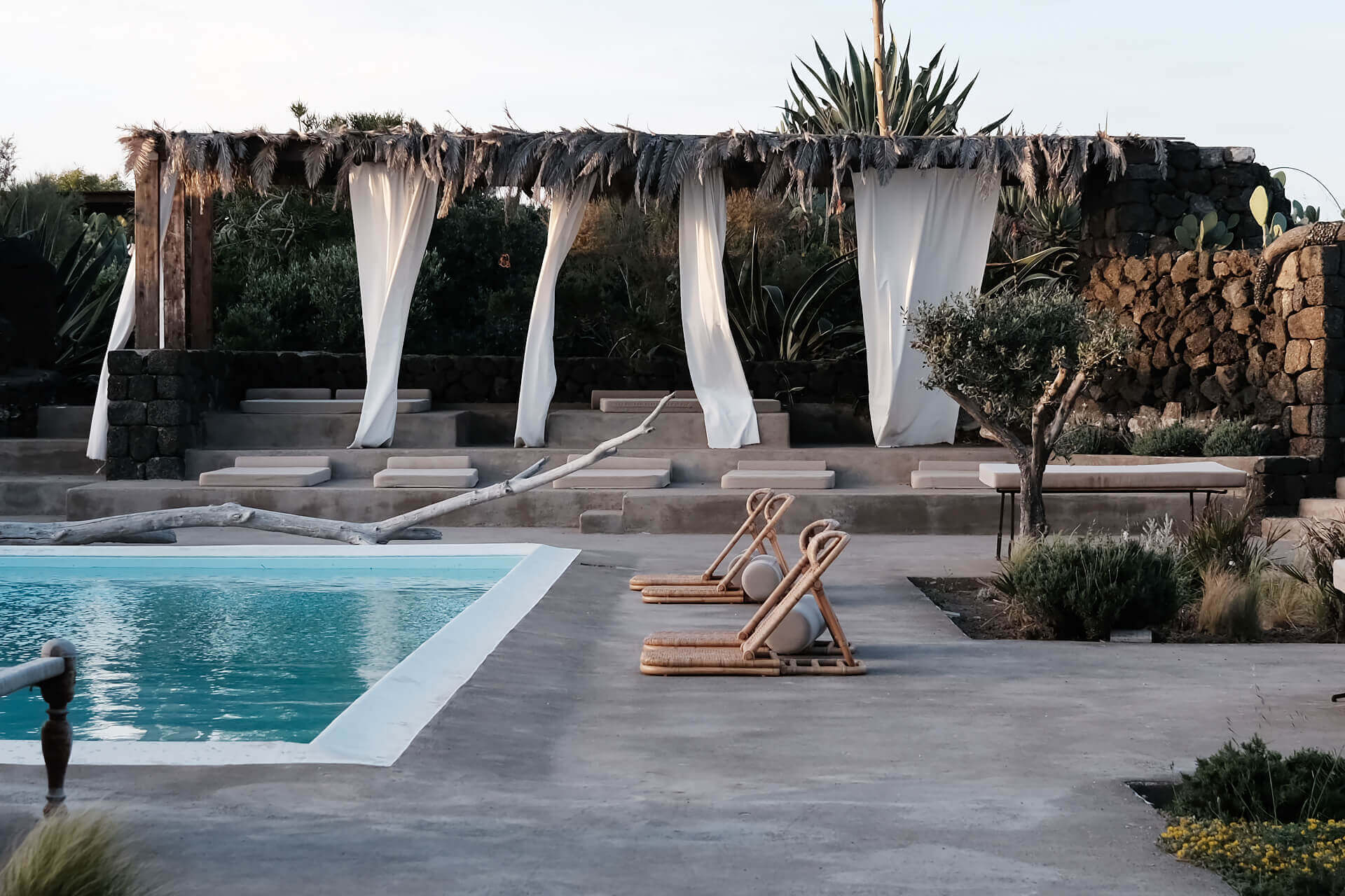 Parco Dei Sesi, Europe — Tiered poolside terrace with turquoise water, wooden loungers, flowing white curtains on pergola, stone walls, Mediterranean plants, evening light.