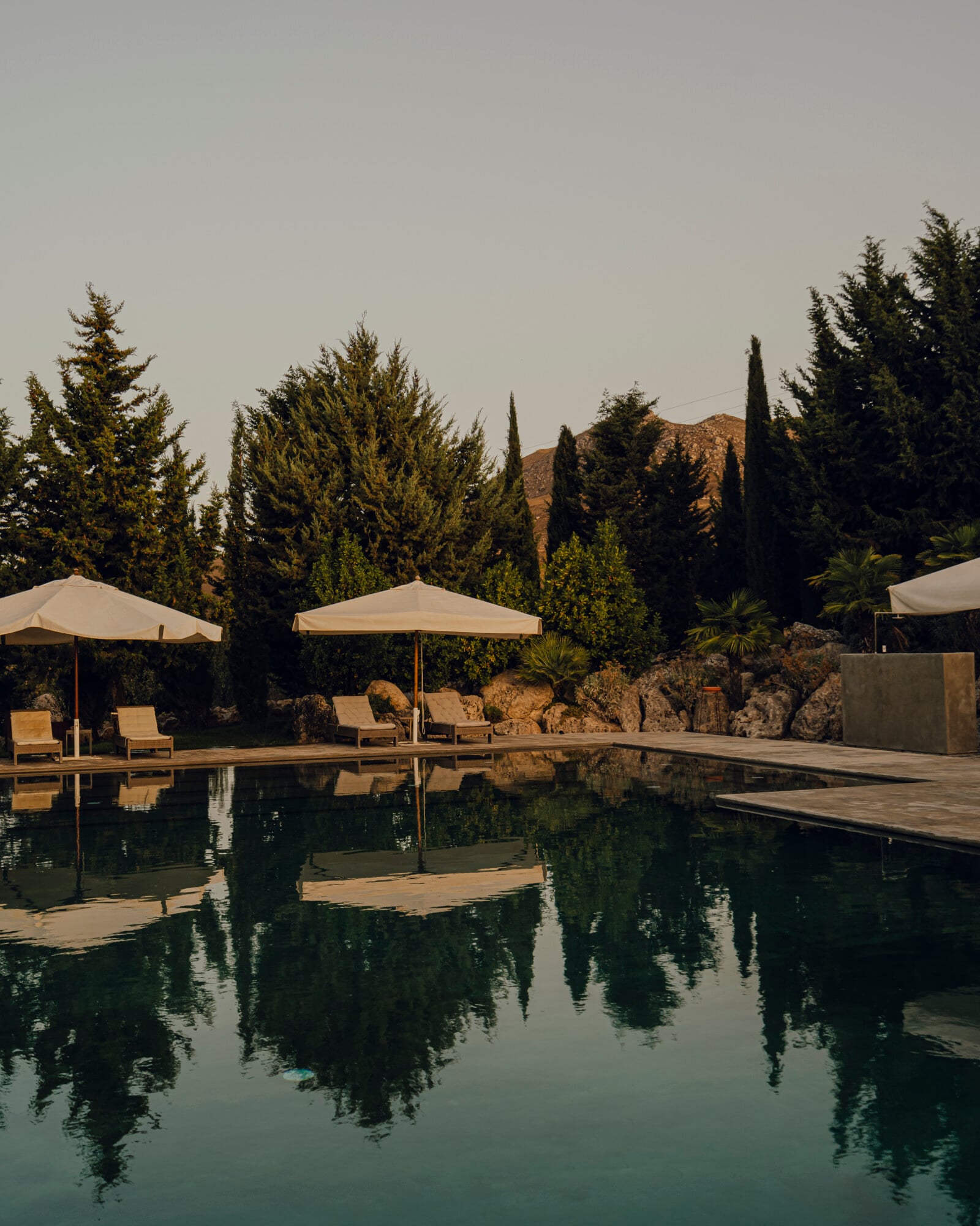 Susafa, Italy — Reflecting pool with cypress trees, stone terraces, cream umbrellas, and arid hillside landscape at sunset.