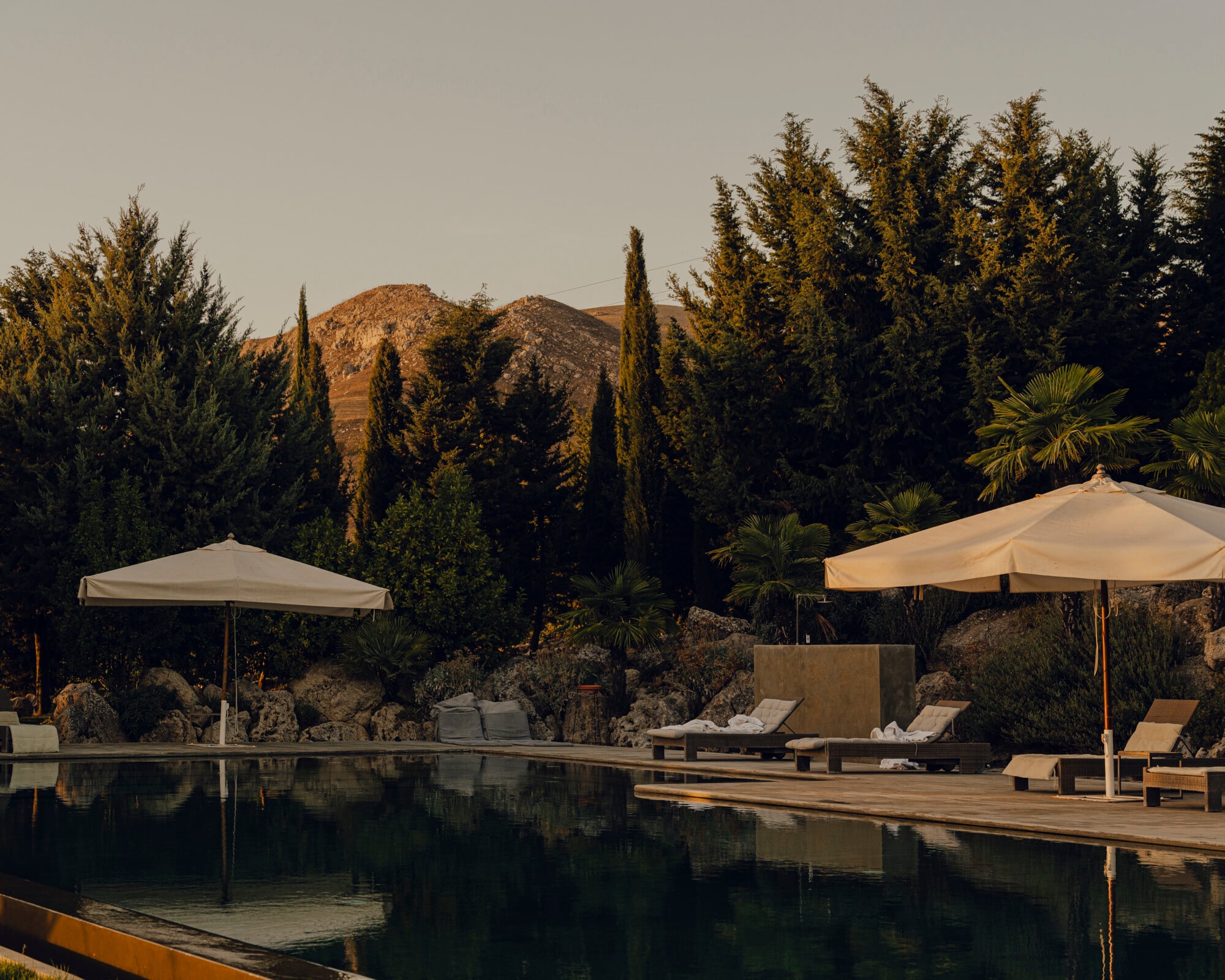 Susafa, Italy — Infinity pool with wooden deck, cream umbrellas, and loungers overlooking cypress trees and rocky hillside at golden hour.