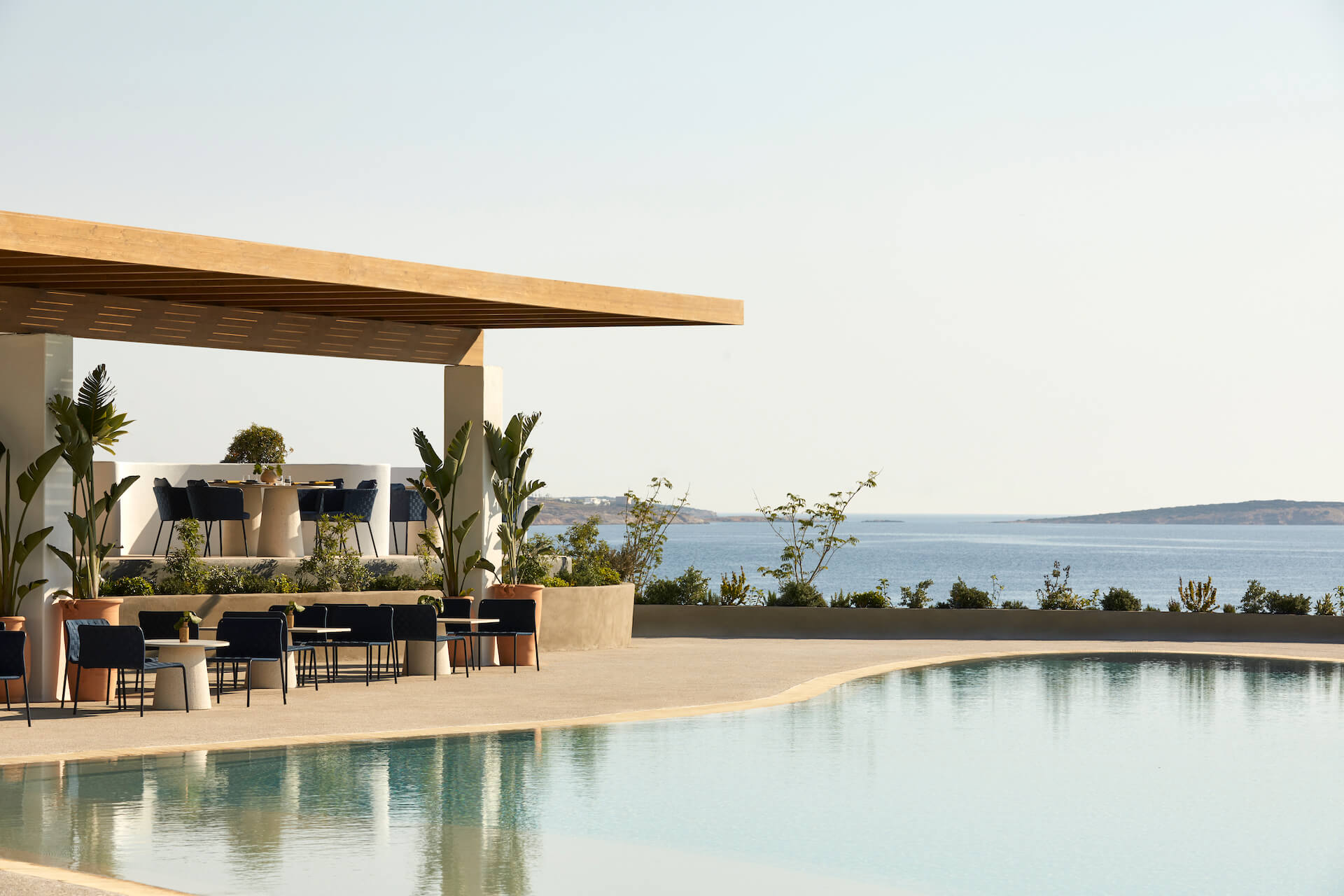 Pārocks, Europe — Modernist poolside pavilion with timber pergola, black seating, potted plants, and Adriatic Sea views at sunset.