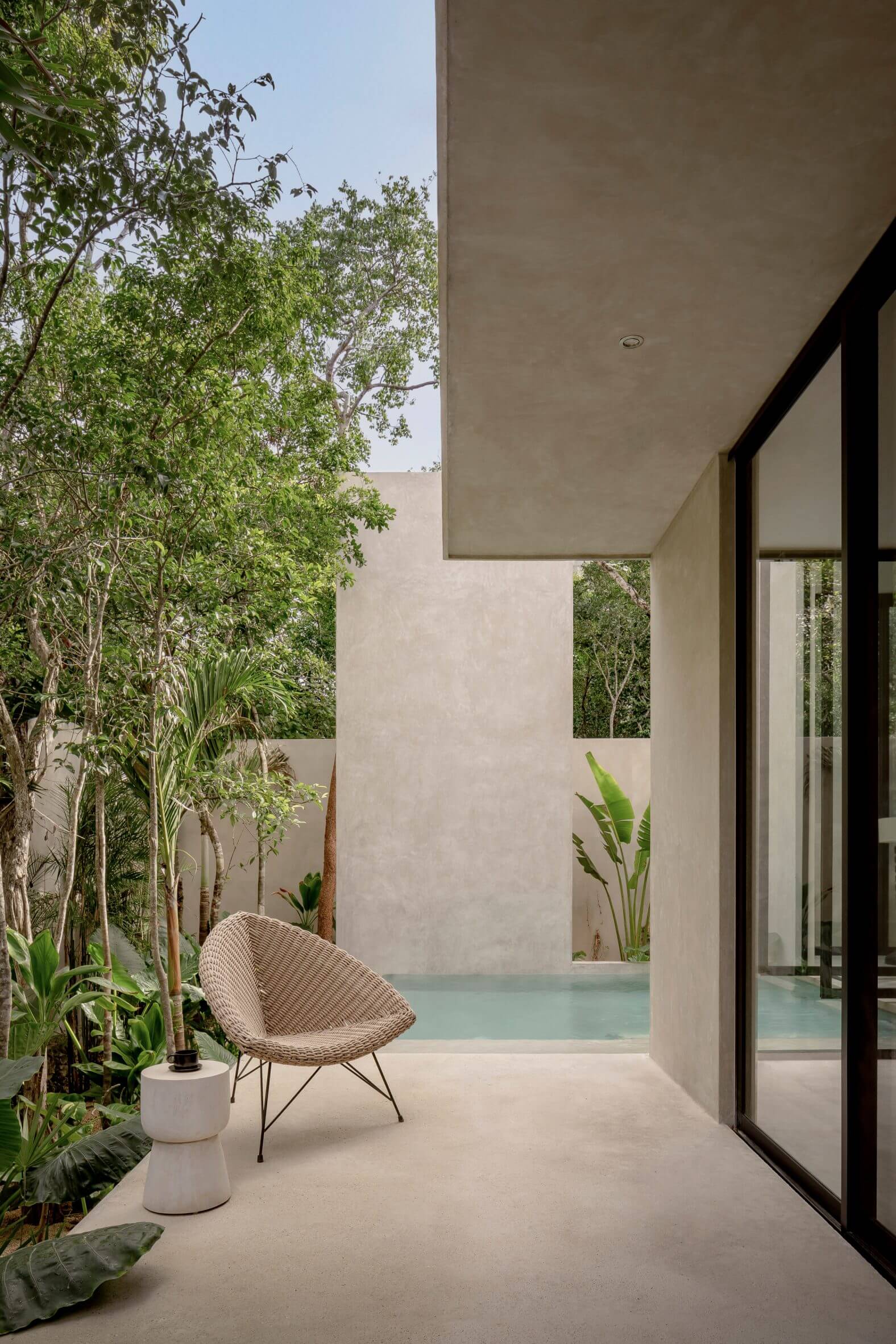 Villa Cava, Mexico — Minimalist courtyard with woven lounge chair, concrete walls, tropical plants, and pool framed by black-framed glass doors.